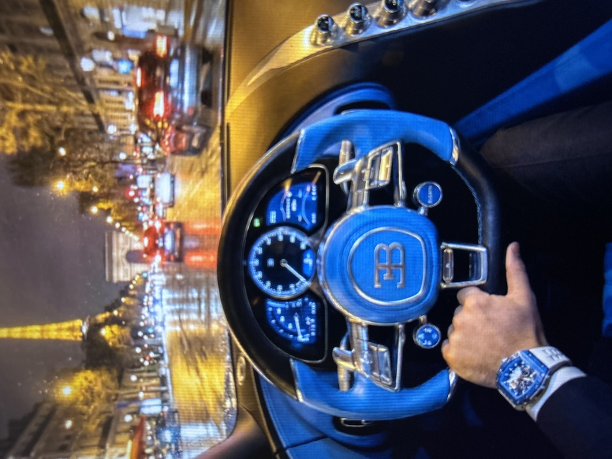 The image shows a first-person perspective from inside a luxury car, likely a Bugatti, as evidenced by the "EB" logo on the steering wheel and a "Bugatti" button on the dashboard. The car is driving on a wet street at night, with reflections of streetlights and traffic on the road. In the background, iconic Parisian landmarks are visible, including the Eiffel Tower and the Arc de Triomphe. The driver's hand, wearing a high-end watch, is resting on the steering wheel. The dashboard features digital displays with blue backlighting. The environment appears to be a city street with ornate buildings and trees lining the road, illuminated by streetlights. The scene suggests a luxurious and high-speed experience in Paris at night.