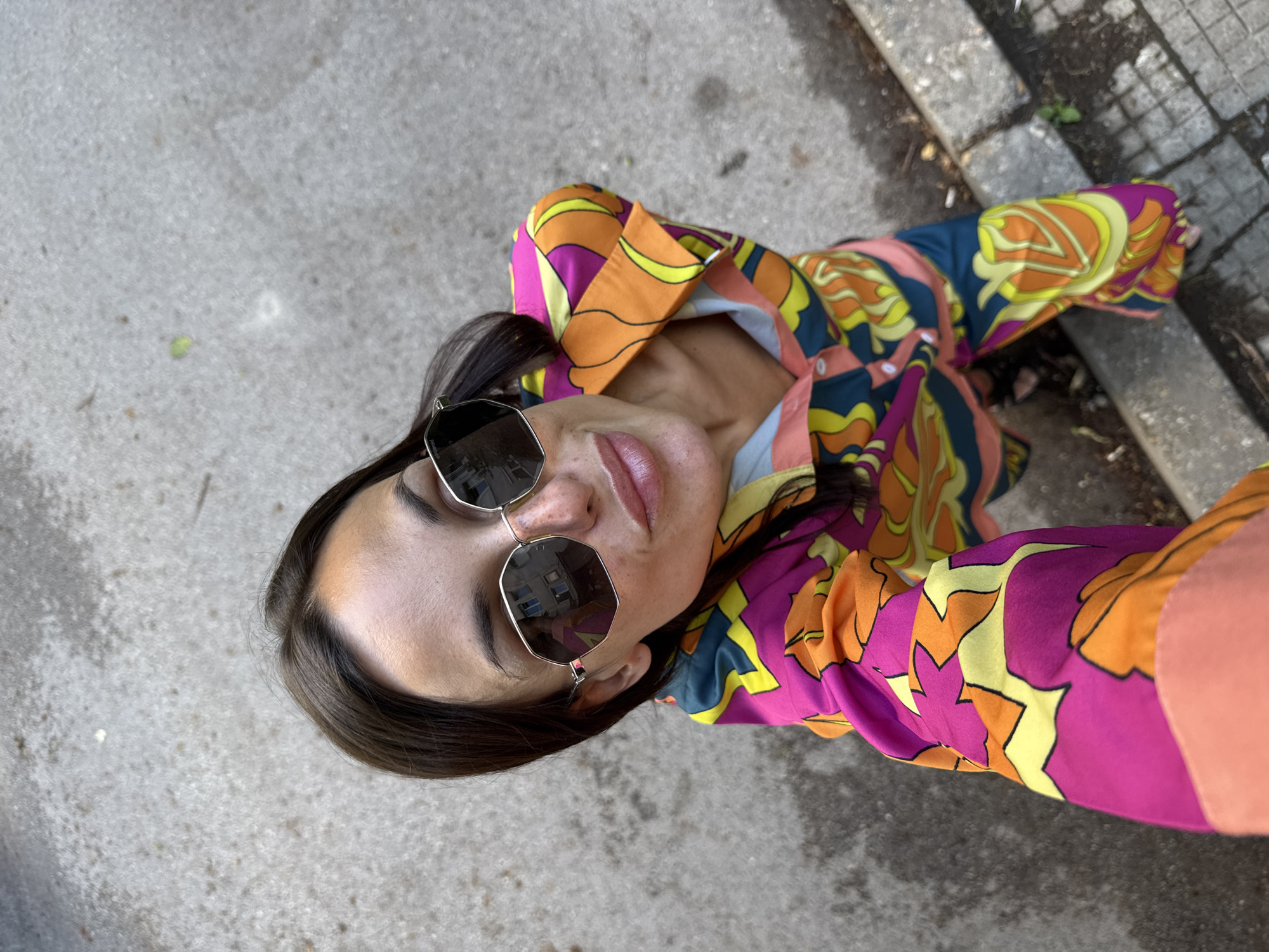 A woman is taking a selfie outdoors on a paved surface in the city of Pleven, Bulgaria. She is wearing a brightly colored, patterned shirt and pants. She has dark hair and is wearing sunglasses. The ground is grey and cracked, with some debris and a curb visible. The background is out of focus.