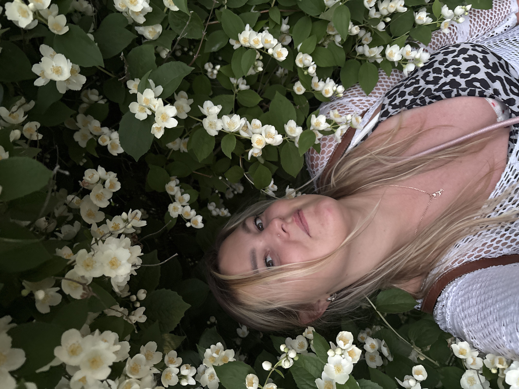 A woman with blonde hair and a white lace top is positioned in the center of the frame, partially obscured by white jasmine flowers and green leaves. She is looking towards the camera with a slight smile. A leopard-print bikini top is visible beneath her lace top. A thin pink strap extends from her shoulder across her chest. Her right ear is adorned with a small silver hoop earring. She wears a delicate rose gold necklace with a small pendant. The background consists of dense foliage of jasmine flowers and leaves. The scene is outdoors, with soft natural lighting. The environment suggests a garden or park setting. It is not possible to determine the specific location within Chaikovsky, Russia, from the provided image.