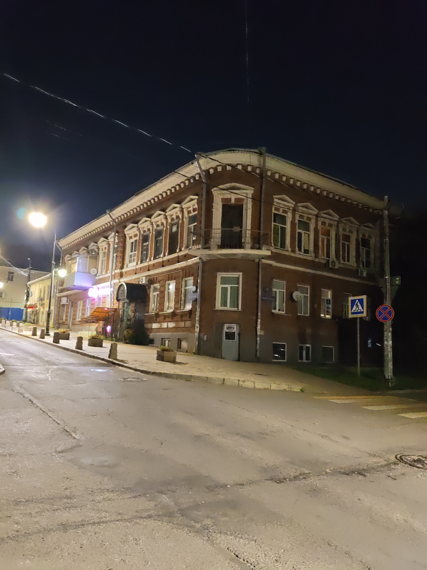 The image depicts a street scene at night in Rostov-on-Don, Russia. A multi-story brick building with ornate window frames and a balcony is the primary subject. The building appears to be a mixed-use structure, with businesses visible on the ground floor, indicated by illuminated signage and doors. A streetlamp illuminates the road, which is wet, suggesting recent rain or damp conditions. The street is lined with planters and bollards. Traffic signs, including a pedestrian crossing sign and a no parking sign, are visible. In the background, other buildings and a dark sky are present. There are no people visible in the image. The overall impression is of a quiet urban street at night.