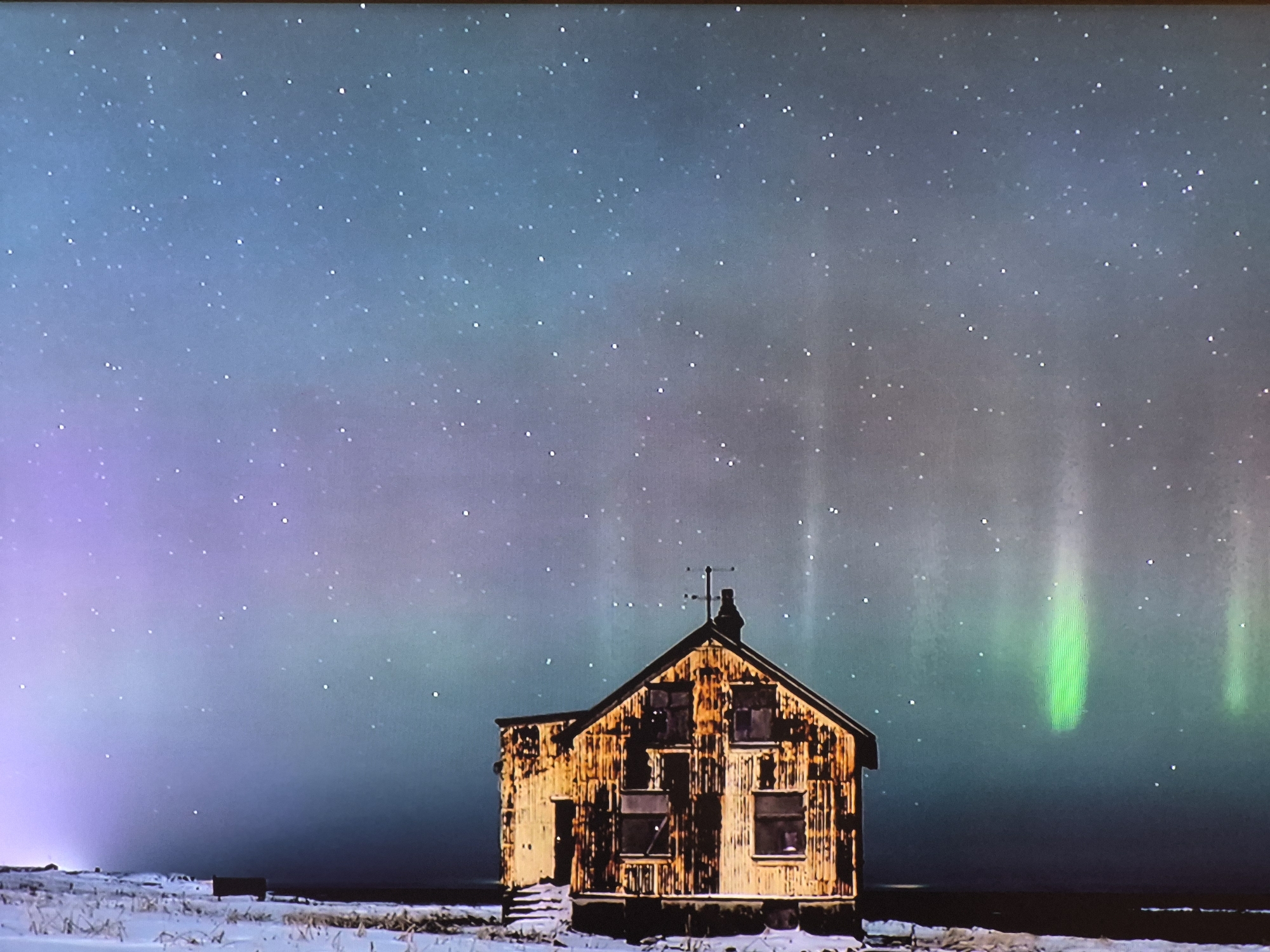 An isolated, weathered, one-story building stands in a snow-covered landscape under a night sky displaying the aurora borealis. The building is characterized by its aged, orange, and brown exterior with visible window frames and a small chimney. The aurora appears as a series of green and purple streaks across the sky, punctuated by stars. The ground is white with patches of snow, and the horizon is dark. The location is not Yerevan, Armenia.