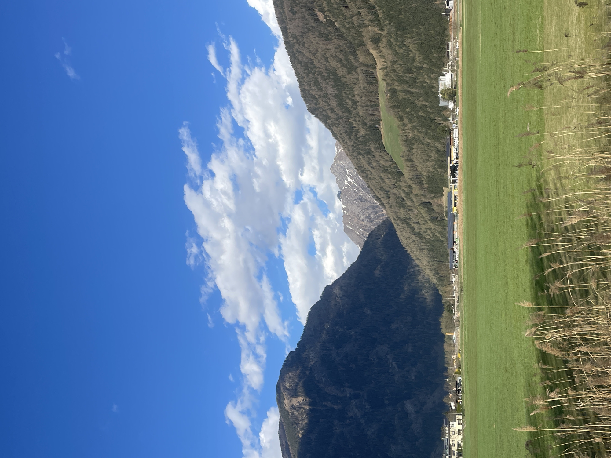 The image showcases a vibrant landscape in Sterzing-Vipiteno, Italy. Dominated by a clear blue sky with scattered white clouds on the left side of the image, it contrasts with the dark, forested mountains that fill the center. A green field stretches along the right edge, bordered by tall, dry grasses. A small settlement is nestled between the mountains and the field, hinting at human habitation within this natural setting.