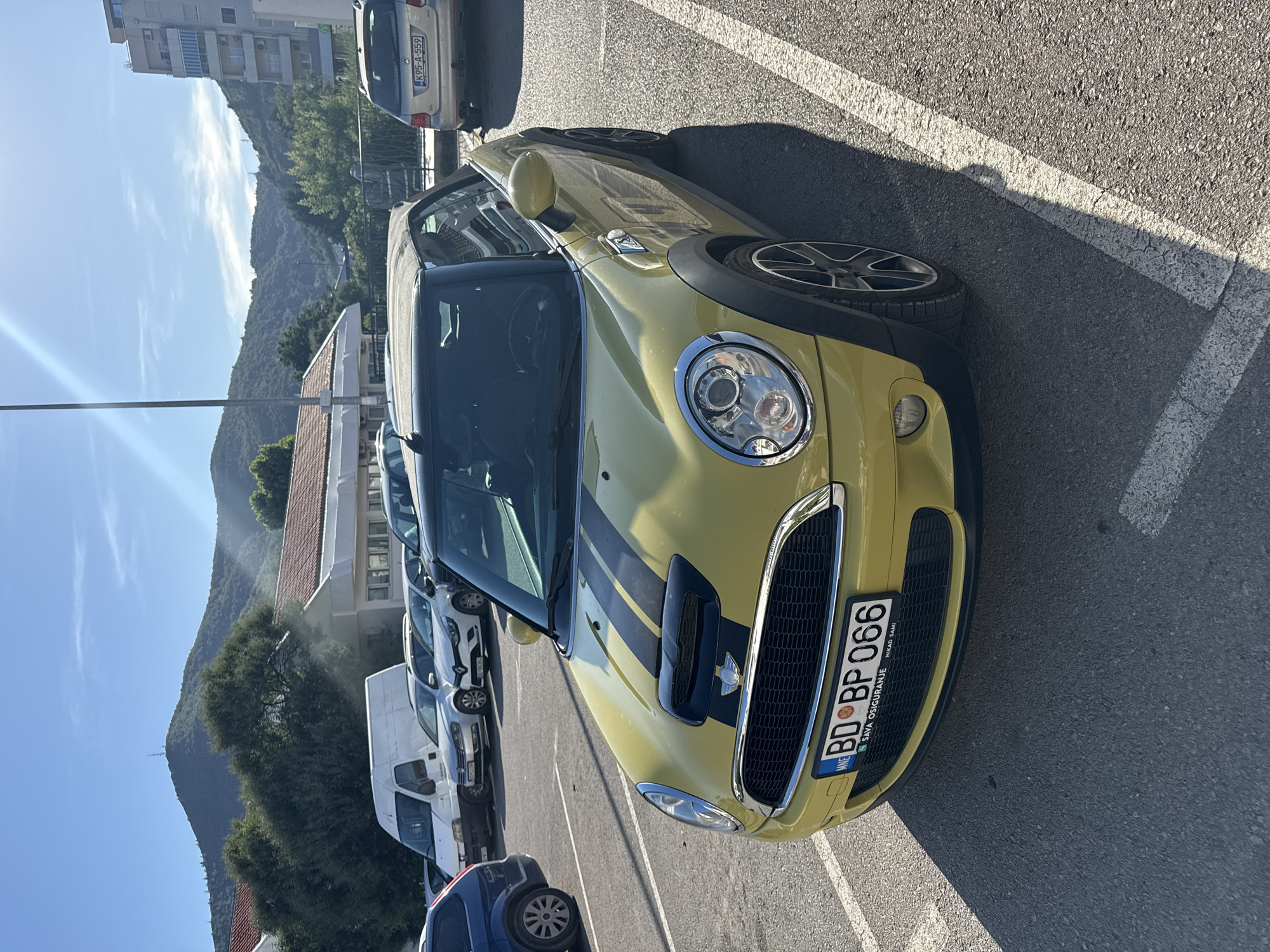 A yellow Mini Cooper convertible is parked in an outdoor parking lot in Budva, Montenegro. The car has black racing stripes on the hood and a black soft top. Its license plate reads "BD BP 066". In the background, there are other cars, buildings, trees, and mountains under a clear blue sky. The sun is shining, casting shadows on the ground.