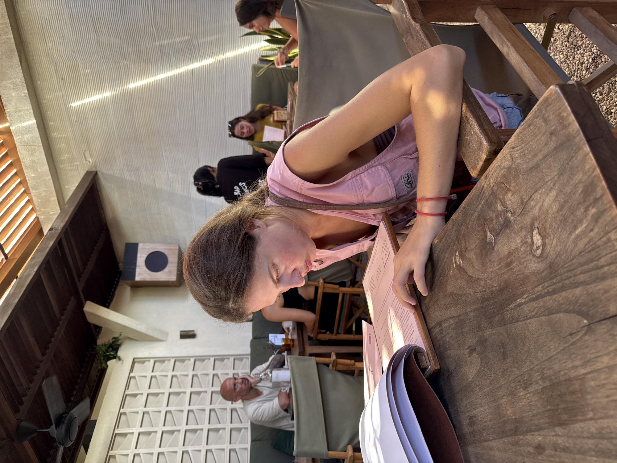 The image shows a woman looking at a menu at a restaurant in Kuta Selatan, Indonesia. She is wearing a pink vest and is sitting at a wooden table. Other people can be seen in the background, including another woman looking at a menu and a man sitting at a table. The restaurant has a partially enclosed roof, and the lighting is bright. A ceiling fan is visible, as well as a speaker on the wall.