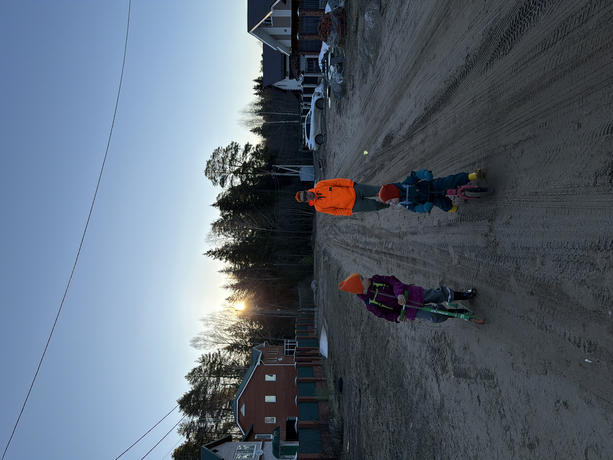 The image depicts a street scene with an unpaved road surface marked with tire tracks. Two children on scooters and an adult are walking along the road. One child is wearing a purple jacket and an orange hat. The other child is wearing a blue jacket and a red hat. The adult is wearing an orange jacket. On either side of the road are houses, some under construction, and trees. The sky is light blue, indicating either early morning or late afternoon. Overhead, power lines stretch across the sky. Given the architecture, general environment, and information provided, the location appears to be in the city of Chaikovsky, Russia.