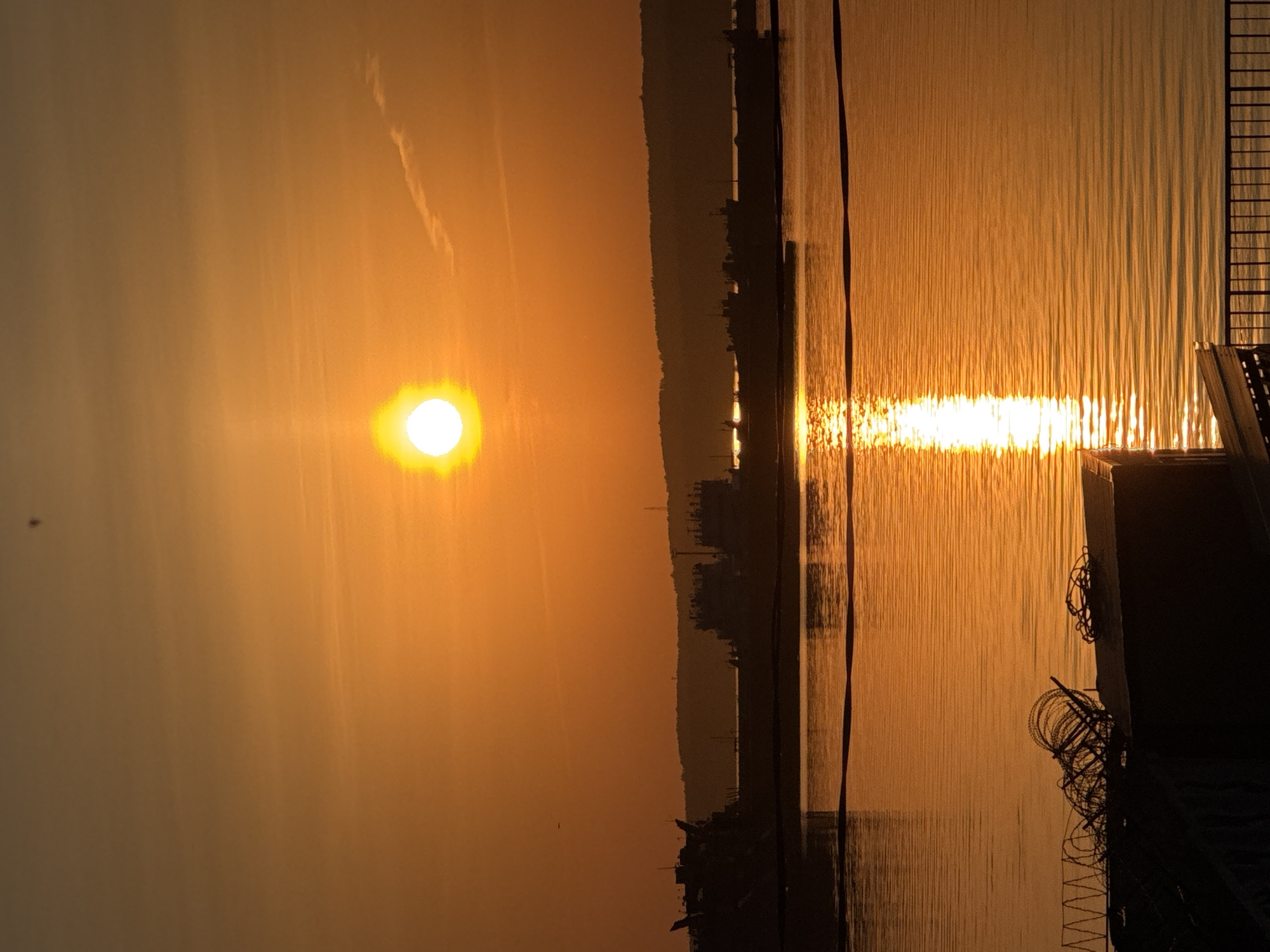 The image shows a sunset scene in Chaikovsky, Russia. The sun is prominently visible in the sky, with a strong reflection on the water. A coastline with silhouette structures that may be buildings or trees is visible on the horizon. In the foreground, parts of a structure (possibly a dock or pier) are visible, with some wire or rope detail present. The overall lighting is warm, due to the sunset.