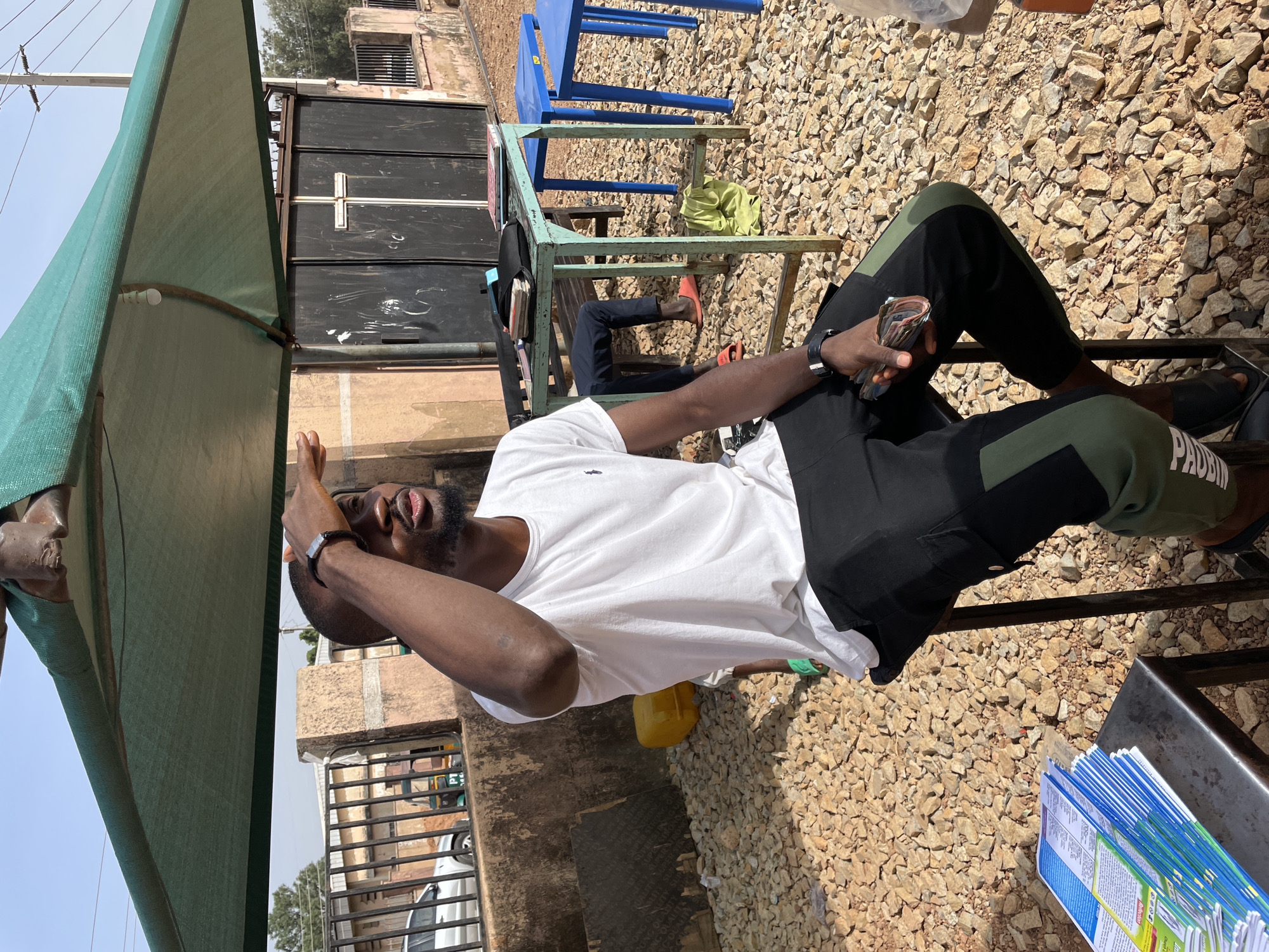The image shows a man seated on a metal chair outdoors in Jos, Nigeria. He is wearing a white t-shirt, black and green pants, and black sandals. He holds a stack of Naira notes in his hand. His left hand is raised to his face, shielding his eyes from the sun. He has dark skin, a beard, and wears a watch on his left wrist.
The man is under a green awning that provides shade. The ground is covered with gravel. In the background are buildings and what appears to be a gate. A blue plastic chair is visible near a metal structure. Some papers or documents are near the corner. Another person is sitting in the background with only their legs visible.
