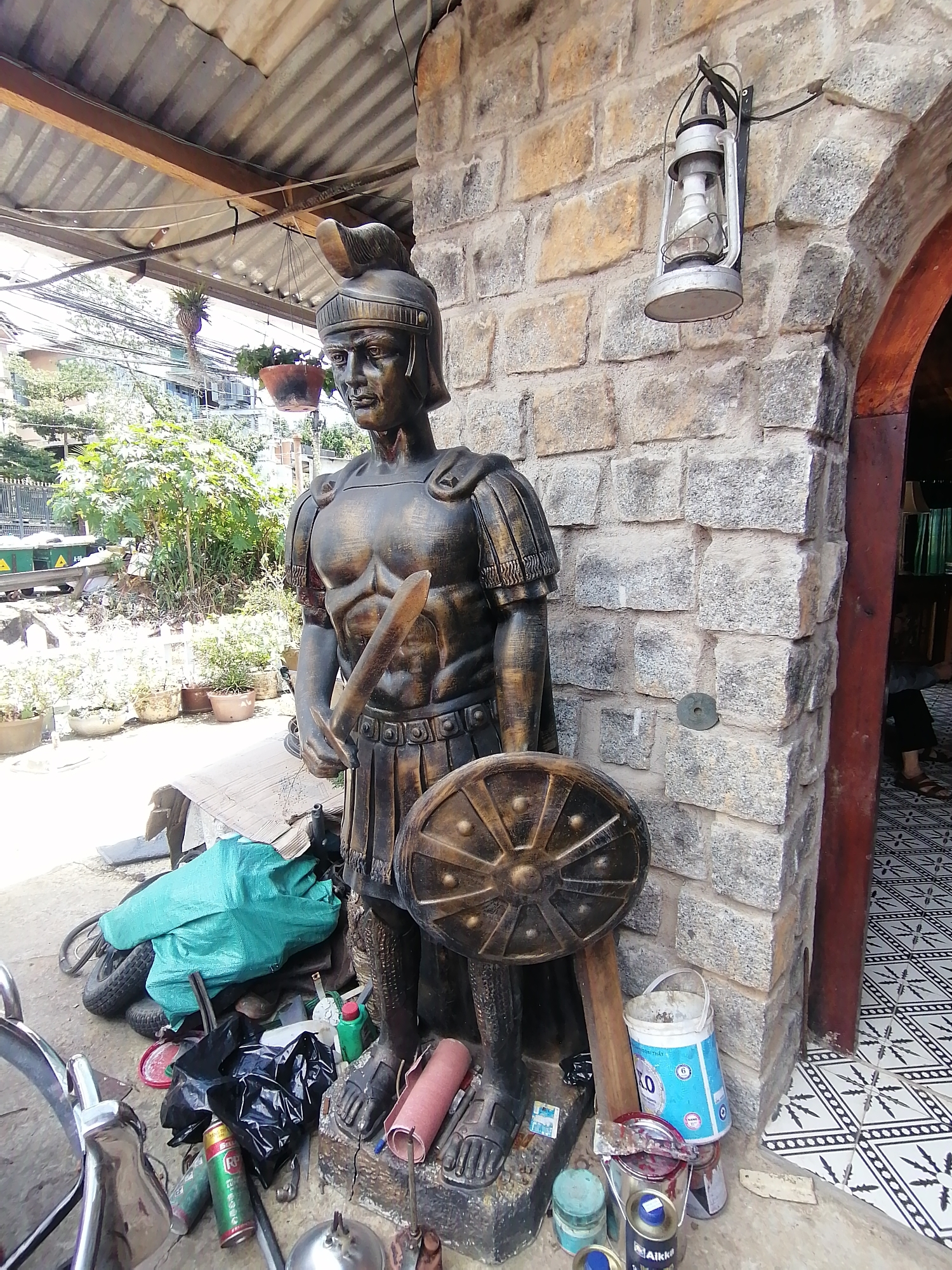 In Da Lat, Vietnam, a life-size statue of a Roman soldier stands guard outside a building. The statue, weathered and dark, is clad in a helmet, chest armor, and a skirt of segmented plates. It holds a sword in its right hand and a round shield in its left. The soldier is positioned against a rough-hewn stone wall, illuminated by a hanging lantern. Surrounding the statue are various objects: discarded tires, a green tarp, black trash bags, paint cans, and other miscellaneous items, suggesting a cluttered outdoor space. Behind the statue, lush greenery and trees are visible, with buildings hinting at the urban environment of Da Lat. The ground is a mix of concrete and patterned tiles.