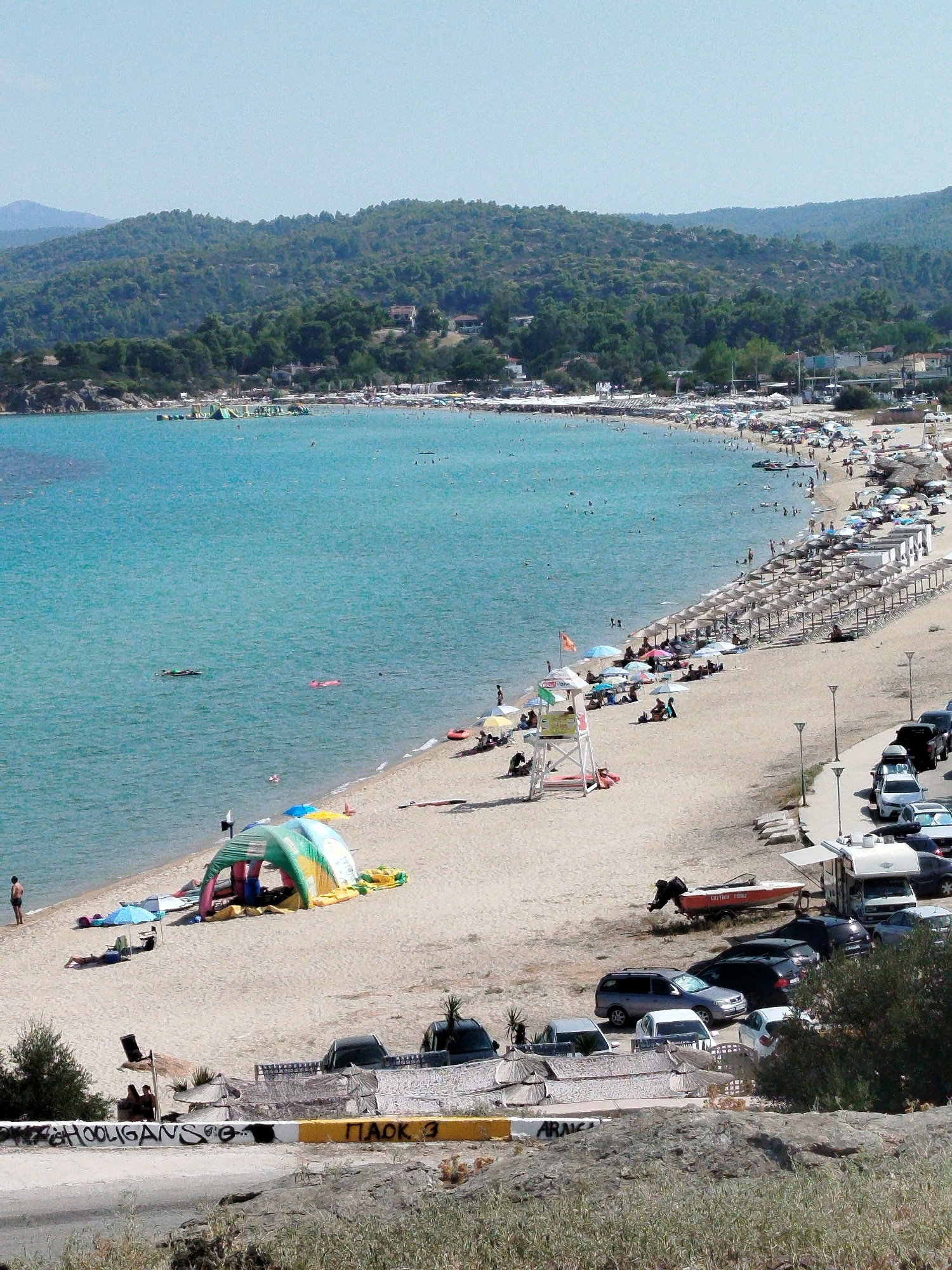 The image depicts a sunny day at a crowded beach in Sithonia Municipal Unit, Greece. The beach is lined with numerous umbrellas and sunbeds, with many people relaxing on the sand and swimming in the clear blue water. A lifeguard tower stands on the beach, and several boats are visible near the shore. There is a parking area with cars and a food truck at the edge of the beach. The background features a lush green hillside with trees and some buildings. A bouncy castle or inflatable water park structure is visible in the shallow water. The overall scene conveys a vibrant and active beach atmosphere.