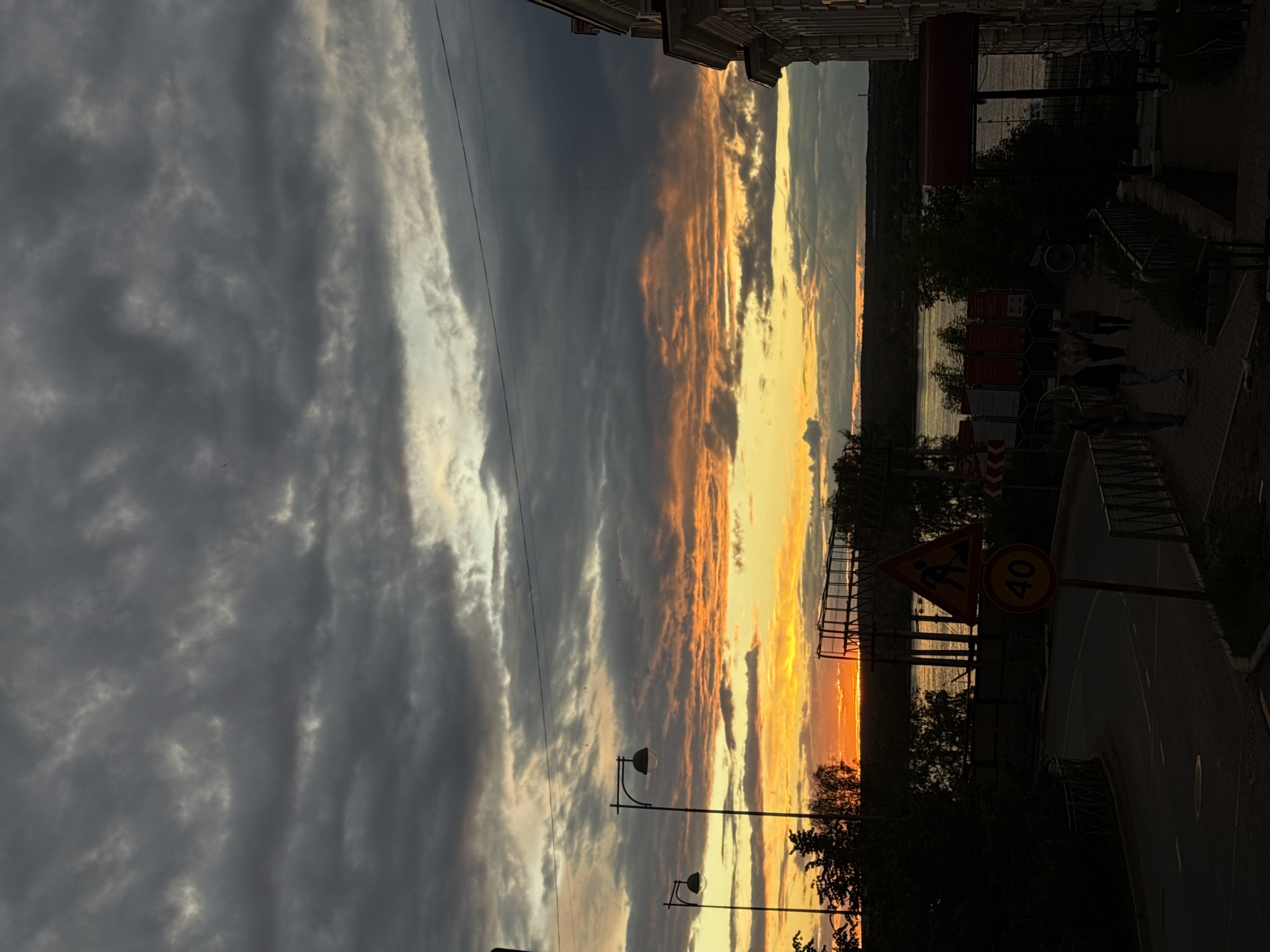 The image shows a sunset in Perm, Russia. The sky is divided between dark gray, striated clouds on the left, and vibrant orange and yellow clouds on the right, reflecting the setting sun. Below the horizon is a body of water, likely the Kama River. Along the embankment, there are street lamps, trees, signs including a "40" speed limit sign and a "men at work" sign, and pedestrian guardrails. People are walking along the embankment. Buildings line the upper edges of the image.