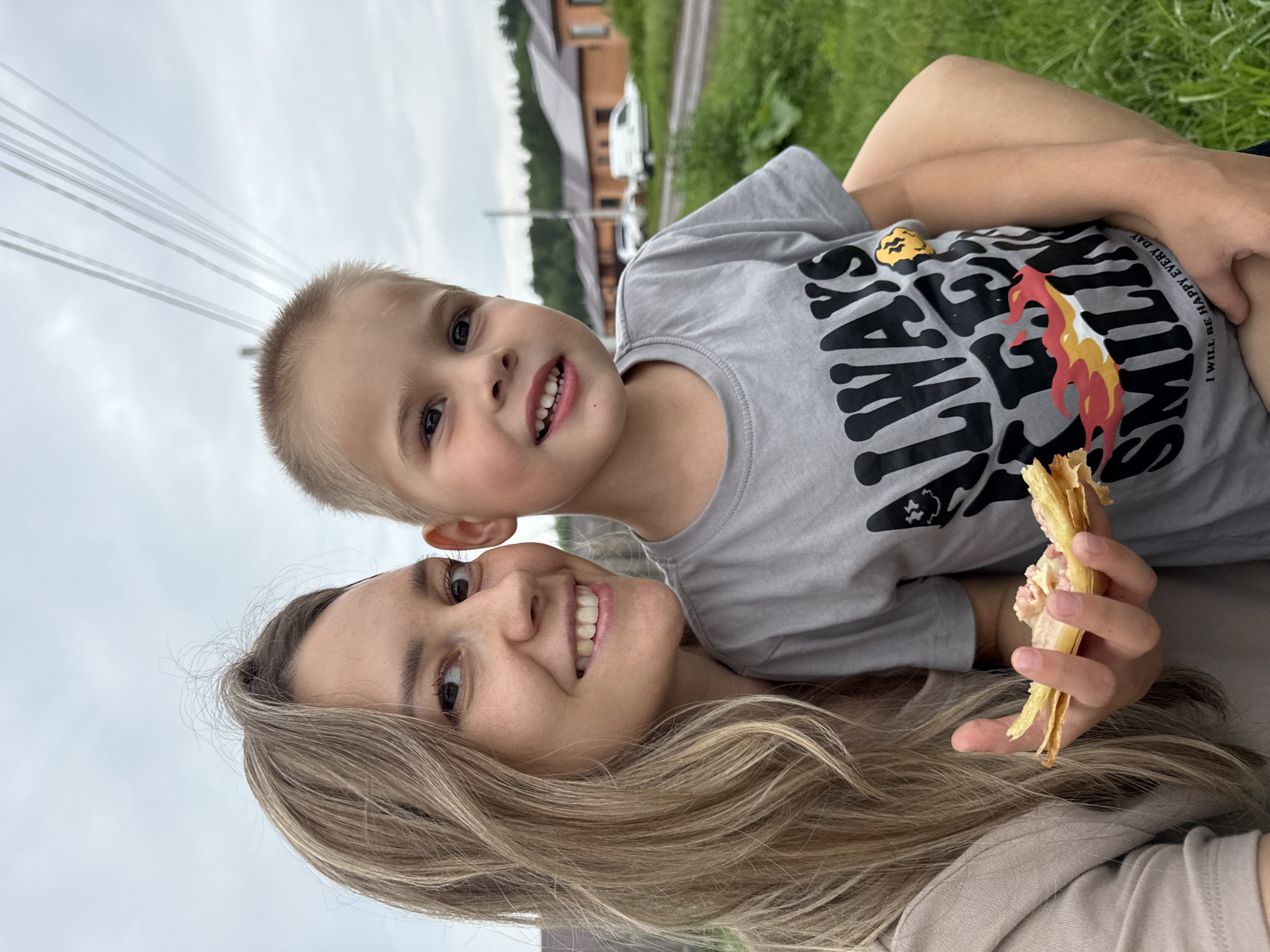 A selfie-style, medium shot shows a woman and a young boy posed outdoors in front of a row of brick houses. The woman, on the left, has long blonde hair and is wearing a tan t-shirt. She is smiling and holding a partially eaten pastry. The boy, on the right, is wearing a grey t-shirt with the words "ALWAYS BE SMILING" and "I WILL BE HAPPY EVERY DAY" printed on it. He is also smiling and has his arm around the woman. The background shows a street with parked cars, green grass, and trees. The sky is overcast. This scene is likely located in the city of Chaikovsky, Russia.