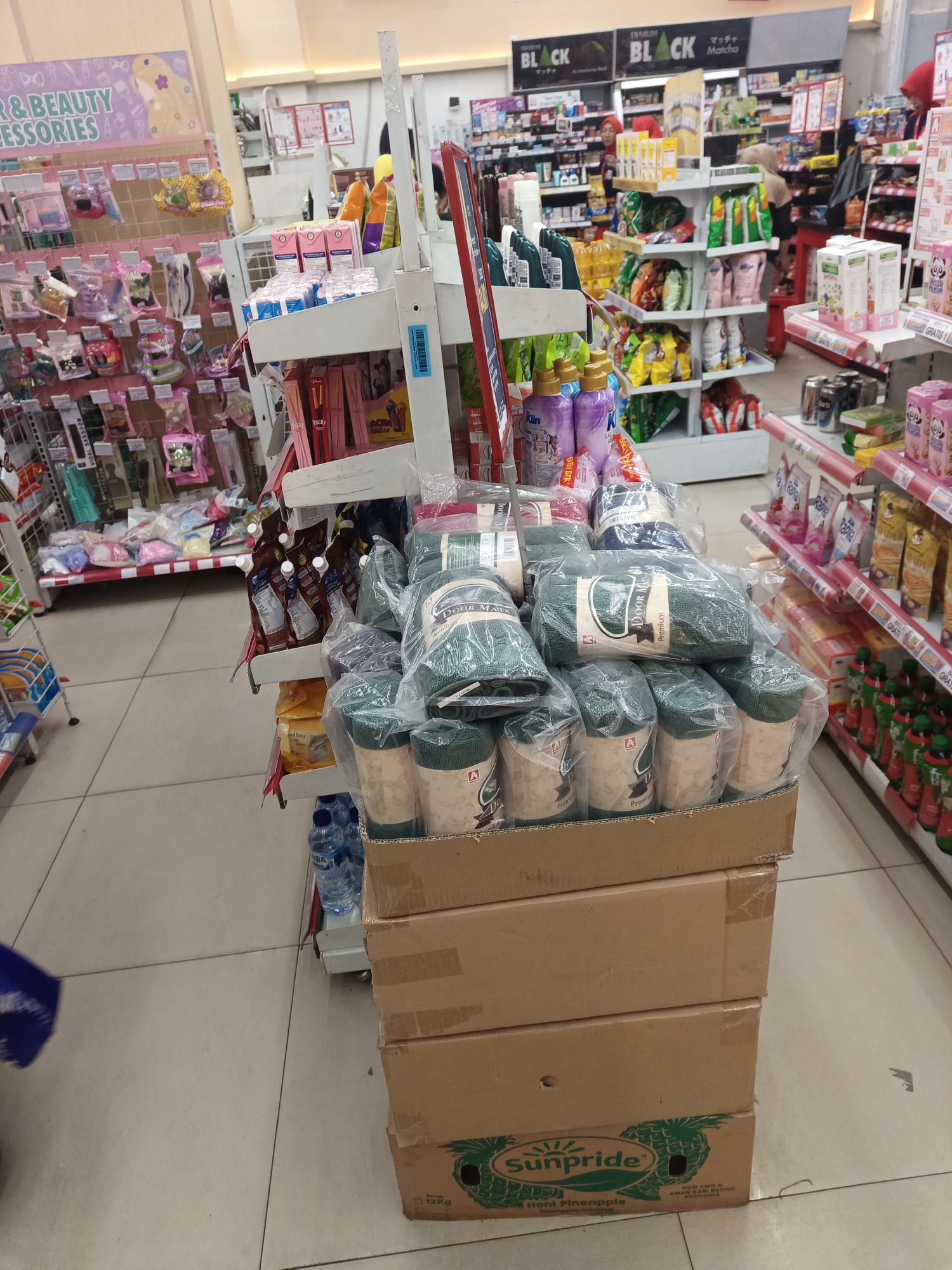 In a well-lit convenience store aisle, likely in North Jakarta, Indonesia, several shelves are stocked with various consumer goods. In the foreground, a display features stacks of boxed items, with the bottom box showing the "Sunpride Honi Pineapple" branding. On top of these boxes are numerous rolled items, some wrapped in plastic and displaying labels like "DOOR MAT Premium."
To the left, a vertical display rack holds an assortment of smaller items, including hair accessories and beauty products. Further down the aisle, more shelves are filled with packaged snacks, beverages, and personal care items. The flooring is tiled in a grid pattern. In the background, other shoppers can be seen browsing the merchandise, and store staff appear to be working. The overall impression is one of a typical retail environment.