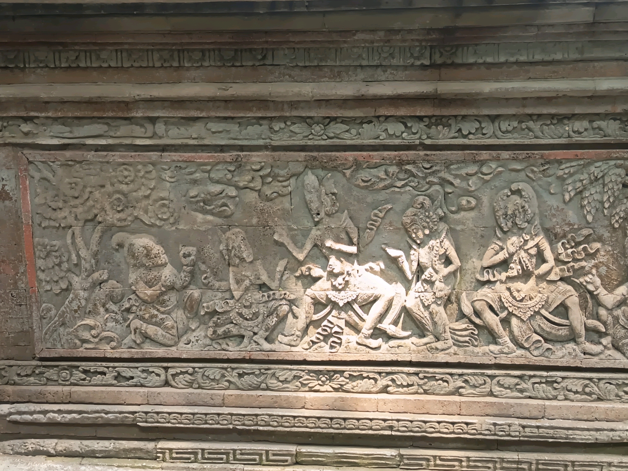 This is a photograph of a stone bas-relief carving on a building located in Girilayu, Indonesia. The carving depicts a scene with multiple figures engaged in various actions. The central figures appear to be human or humanoid, with ornate headdresses and clothing, suggesting a ritualistic or theatrical performance. One figure is seated with legs crossed, while others are standing or in dynamic poses, with some appearing to be interacting with each other. The carving is detailed, showing clothing, jewelry, and facial features. The figures are set against a background that includes stylized natural elements, such as clouds or foliage. The entire relief is framed by decorative borders. The stone itself shows signs of age and weathering.