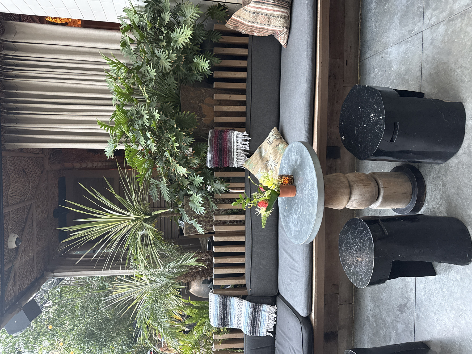 The image shows an outdoor seating area at a restaurant in Los Angeles, California. The seating area features a long, dark gray couch with cushions, a round metal table with a wooden base, and two black stool seats made of what appears to be wood. Throws are draped over the couch, and decorative pillows add to the ambiance. Lush green plants are visible in the background, along with a large beaded curtain. The overall setting suggests a relaxed and natural atmosphere, possibly a patio or garden dining space.