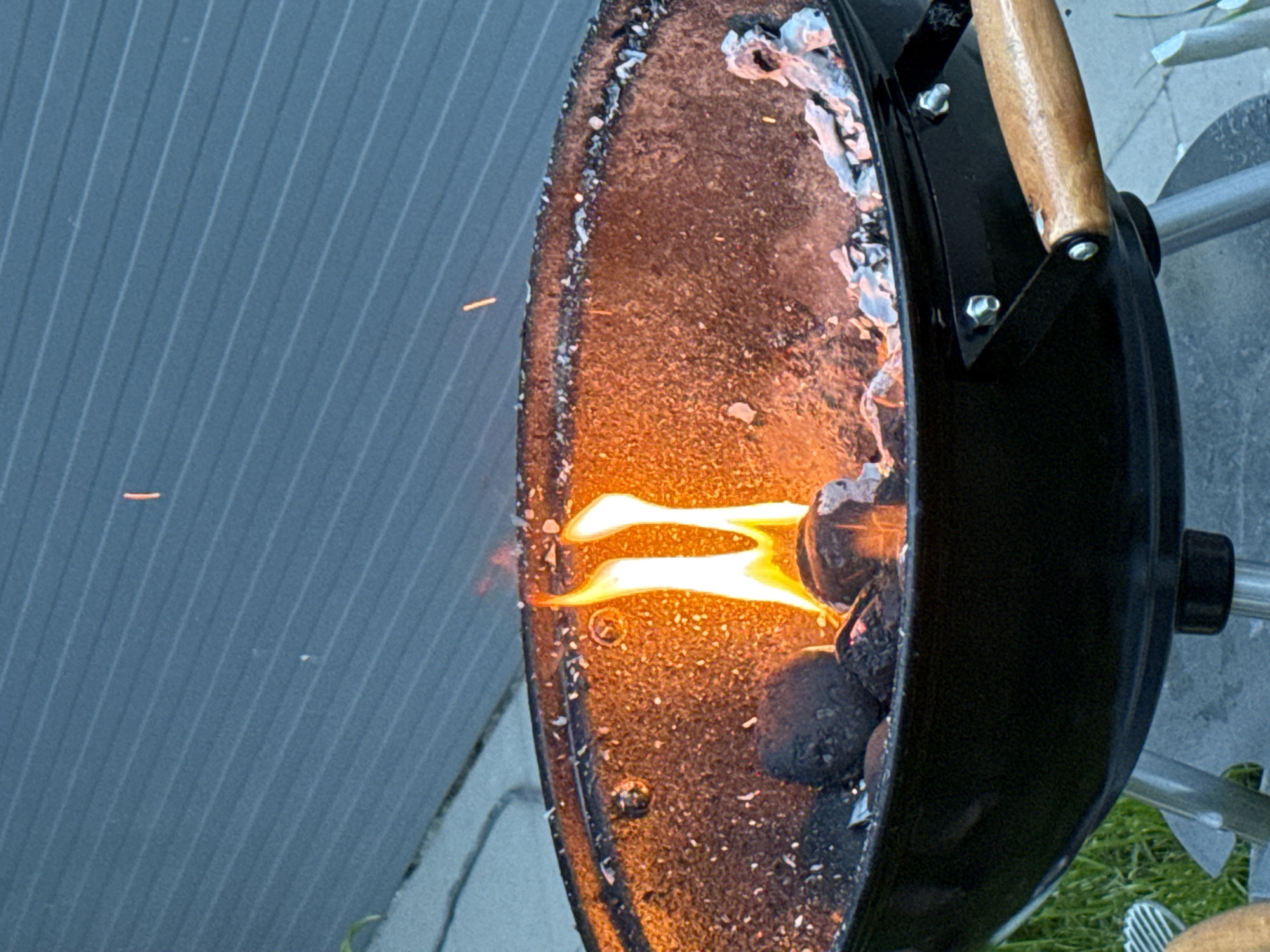 The image shows a charcoal grill with flames emanating from the coals. The grill is black with a wooden handle and is positioned on a grassy surface. In the background, a gray siding of a building is visible. There is no evidence to suggest that this image was taken in Pleven, Bulgaria.