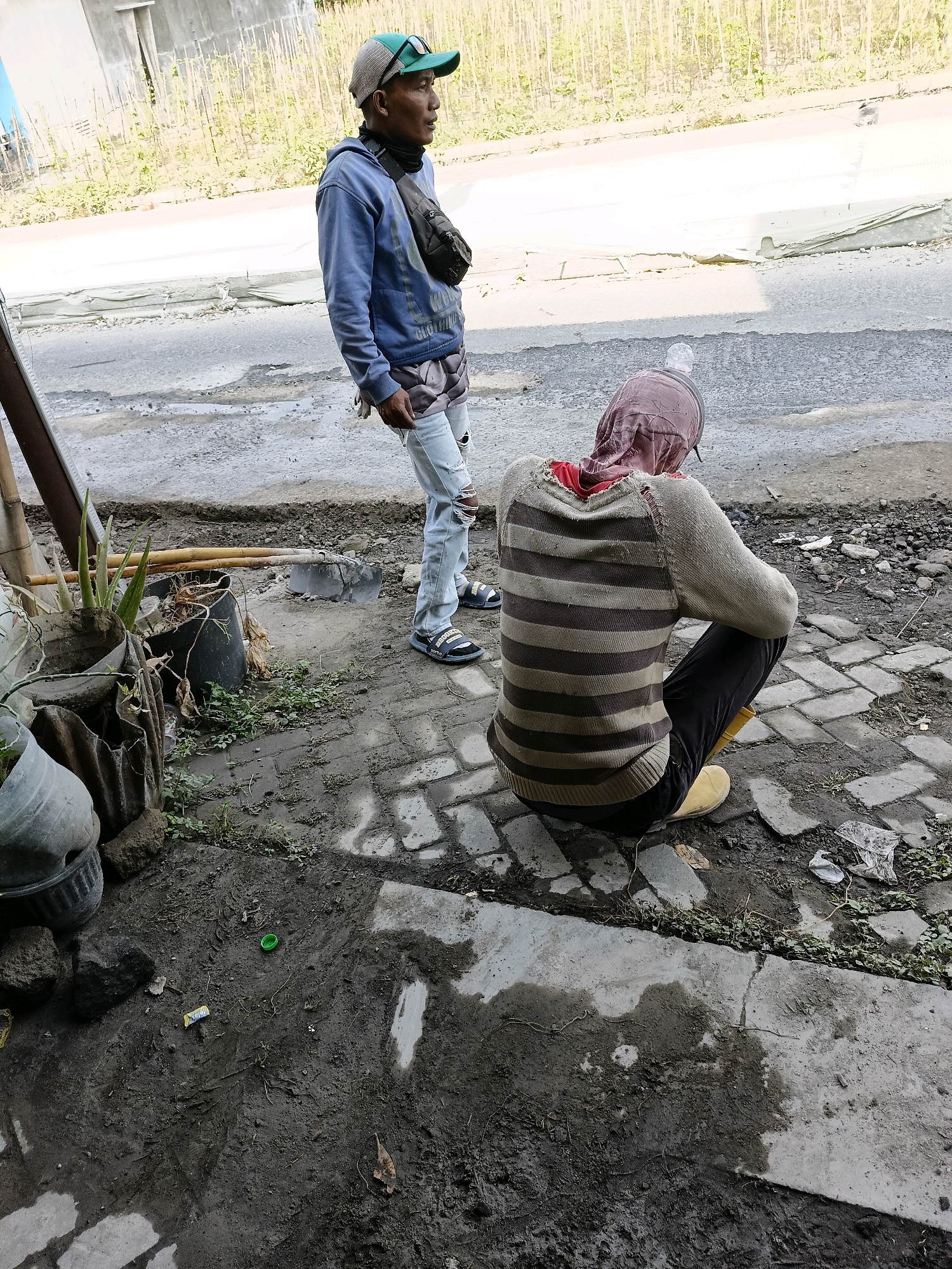 The image shows two individuals in an outdoor setting that appears to be a construction or road work area in Sukorini, Indonesia. The foreground features a muddy ground with cobblestone pavers and scattered debris including a green bottle cap, a wrapper, and a dried leaf. To the left, several potted plants are visible, some in damaged or makeshift containers.
One individual, wearing a blue hooded sweatshirt, ripped light-wash jeans, and a green baseball cap with sunglasses perched on top, stands looking to the right. They have a dark fanny pack worn across their chest. This person is standing on a paved surface that transitions into a road.
The second individual is seated on the cobblestone pavers, facing away from the camera, towards the road. They are wearing a striped long-sleeved sweater with horizontal bands of tan and dark brown, a pink head covering, and yellow boots. Their dark pants are visible as they crouch.
In the background, a road is visible with evidence of repair or construction work, indicated by exposed gravel and what appears to be a partially laid concrete barrier. Beyond the road, a fence made of vertical wooden poles separates the area from a field of tall, dry grass or reeds. The overall environment suggests a sunny day, with harsh lighting creating strong shadows.