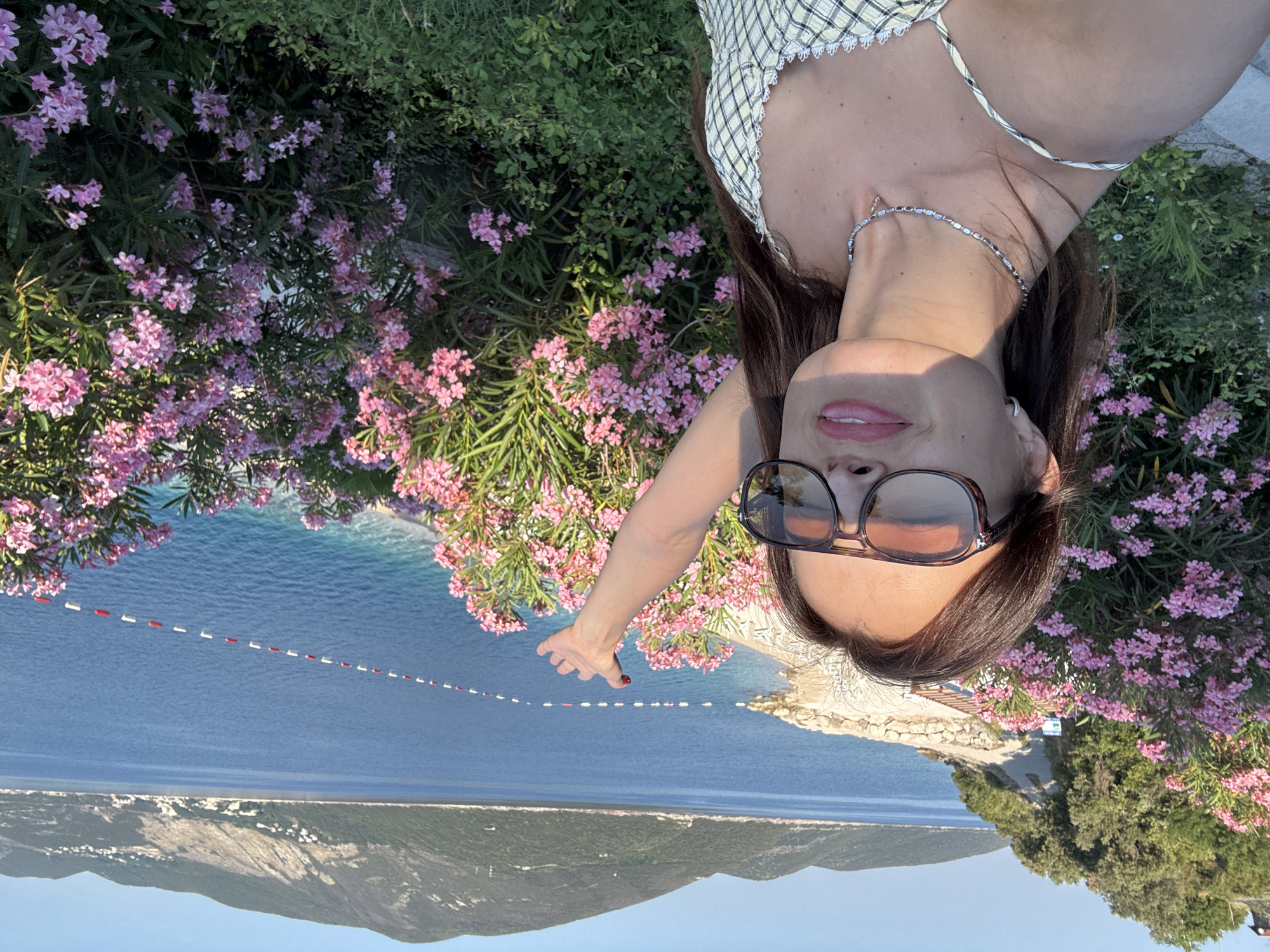 A woman is standing outdoors, in front of a body of water and hills. She is wearing a yellow and white checkered dress, sunglasses, and a necklace. Her arm is raised, and she is looking at the camera. There are pink flowers in the foreground. The sun is shining, and the sky is blue.