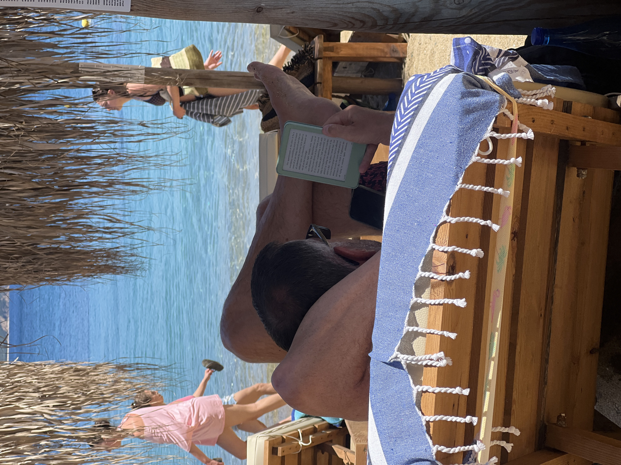 A man is relaxing on a beach lounger in Achladias, Greece, reading an e-reader. The lounger is made of wood and covered with a blue and white striped towel. In the background, two women are walking on the beach near the turquoise water. One woman is wearing a pink dress, and the other is in a patterned bikini. Beach umbrellas provide shade over the loungers. The environment is sunny with clear blue skies.