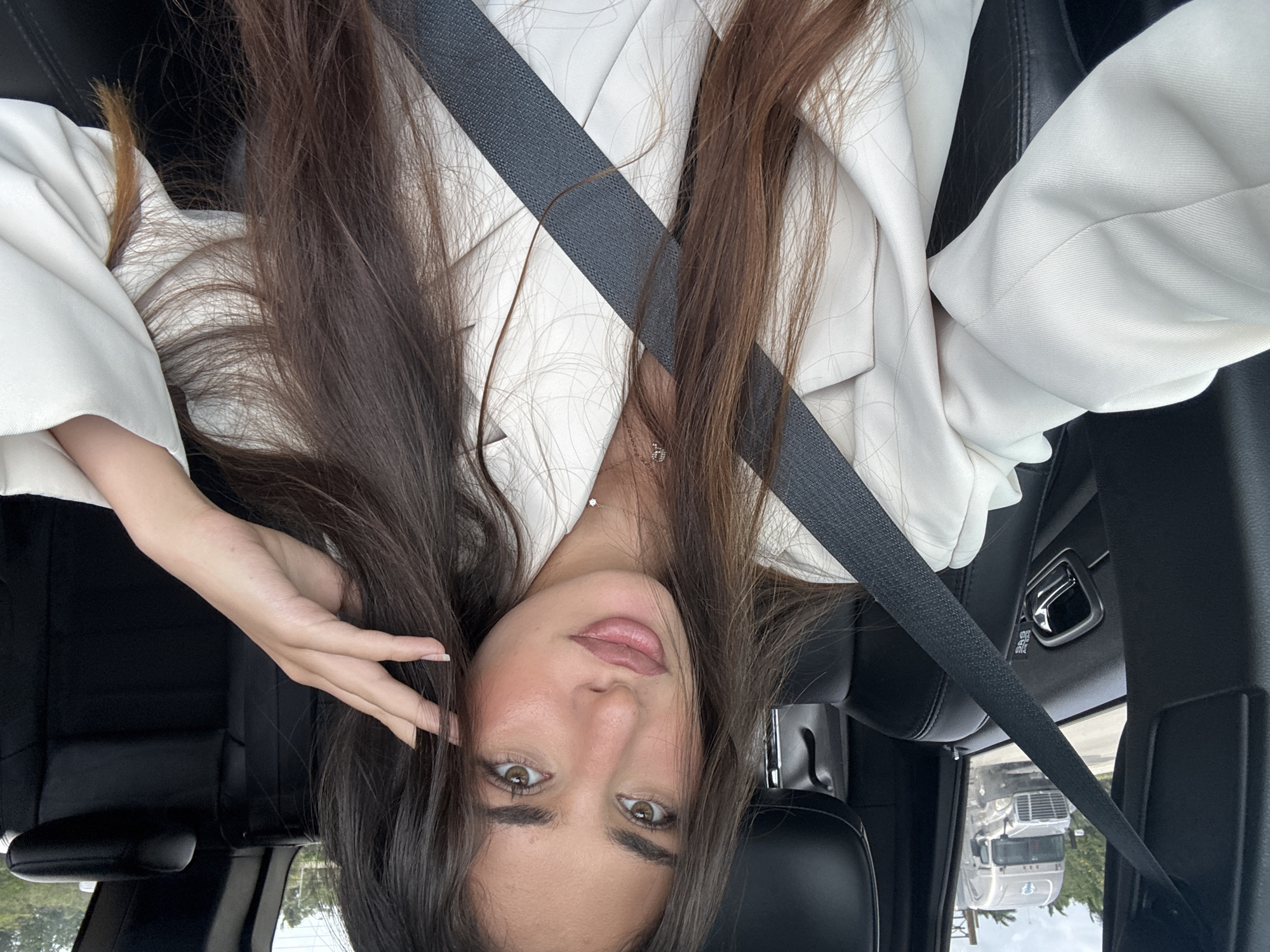 A young woman is in a car wearing a white blazer and a seatbelt. She has brown hair and light brown eyes. A large truck is visible through the window. The location is likely in the United States of America, based on the presence of the truck type and the appearance of the car. The city of Miami, however, cannot be confirmed solely based on the image.