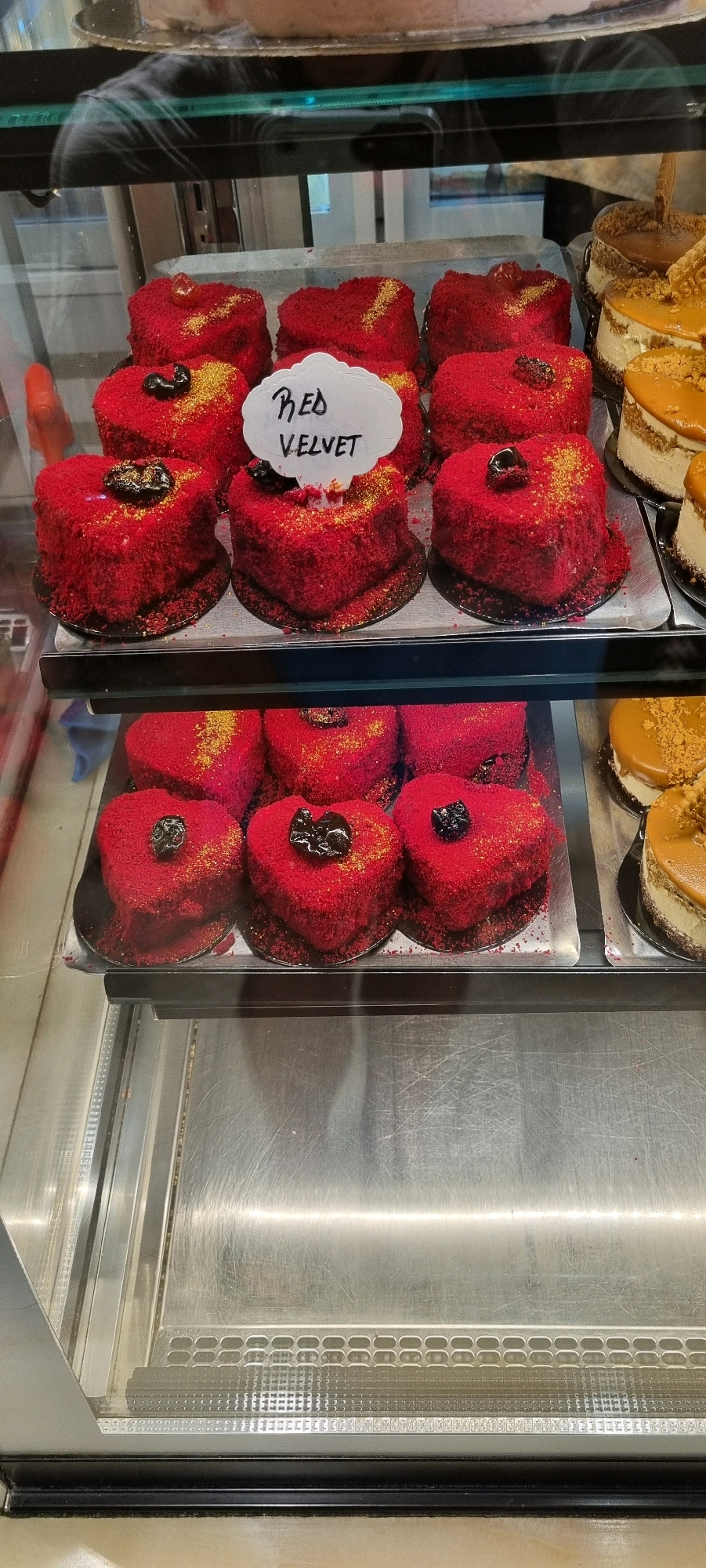 The image shows a display case in a bakery, likely located in Bashkia Durrës, Albania. The display case contains several heart-shaped red velvet cakes, some with gold sprinkles and a dark cherry or fruit topping. A small white sign on one of the cakes reads "Red Velvet". To the right, there are two round cakes with caramel topping and crushed cookies. The cakes are arranged on metal trays within the glass-fronted display case, which appears to be made of stainless steel. The background is blurred but suggests a retail environment.