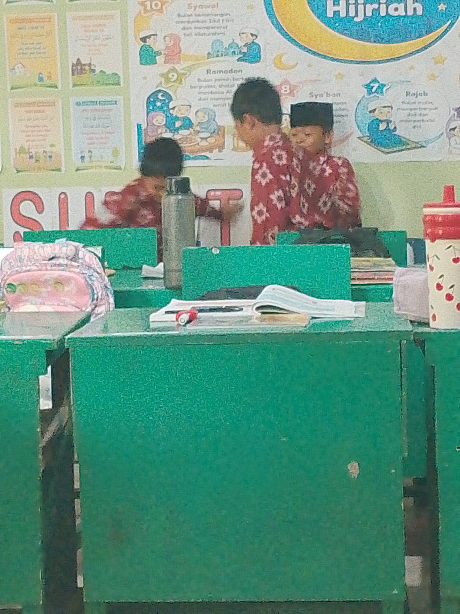 In Tembung, Indonesia, a classroom scene unfolds with two young boys, likely students, standing near green desks. Both boys are wearing traditional Islamic attire. One boy, on the left, is wearing a red shirt with white floral patterns and a black peci (cap). The other boy, to his right, is also dressed in a similar red shirt with white patterns and a black peci. In front of them are several green desks. On one desk, a pink backpack with a patterned cover is visible. Another desk holds a water bottle with a red top and a cherry pattern, next to it a purple cloth. A partially opened book lies on a desk in the foreground. The background displays educational posters with Islamic themes, including a large graphic of a crescent moon and the text "Hijriah." The posters depict illustrations related to Islamic months and traditions, such as people praying and gathering. The overall impression is a classroom setting during an Islamic educational activity or lesson.