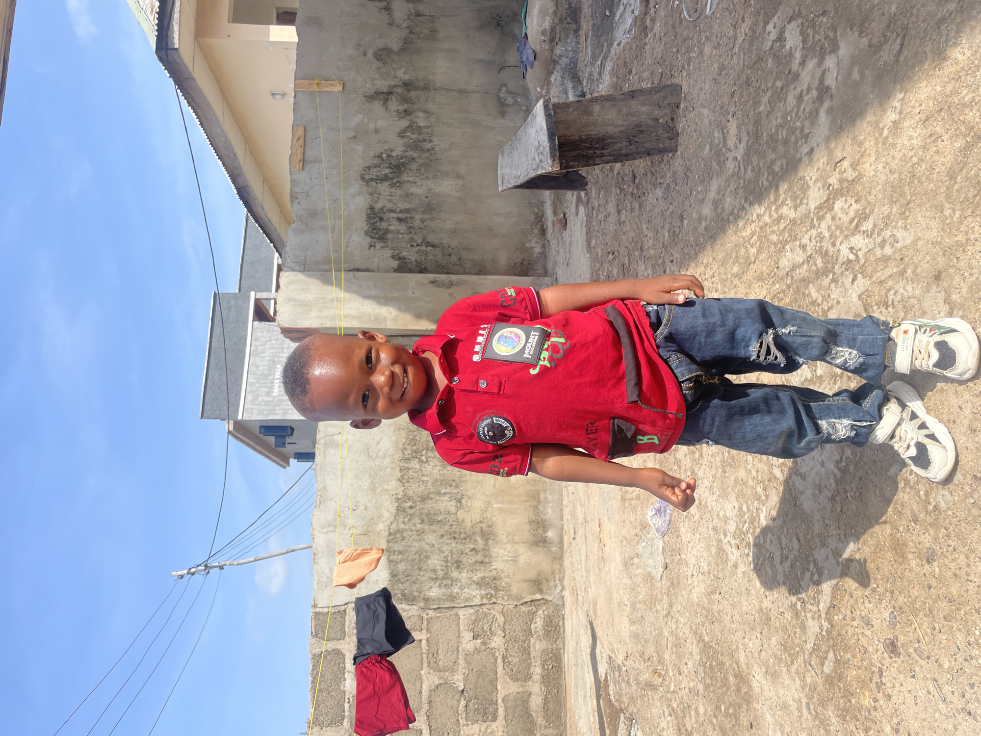 A young boy stands on a concrete surface in Lokoja, Nigeria. He wears a red collared shirt, dark blue jeans with rips, and white sneakers. Behind him is a wall, a hanging clothesline with three pieces of clothing, and part of a building. A small wooden bench sits against the wall. The sky is visible in the background.