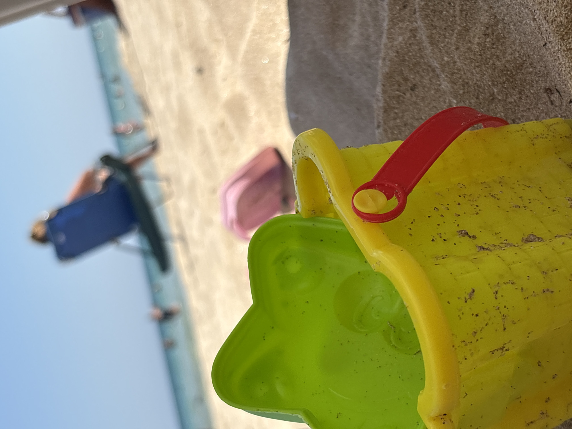 The image depicts a close-up view of a yellow plastic bucket with a red handle and a green sand mold on a sandy beach. In the background, out of focus, are the sea, people swimming and relaxing on beach chairs, and a clear blue sky. The scene is set on the beach in Primorsko, Bulgaria.