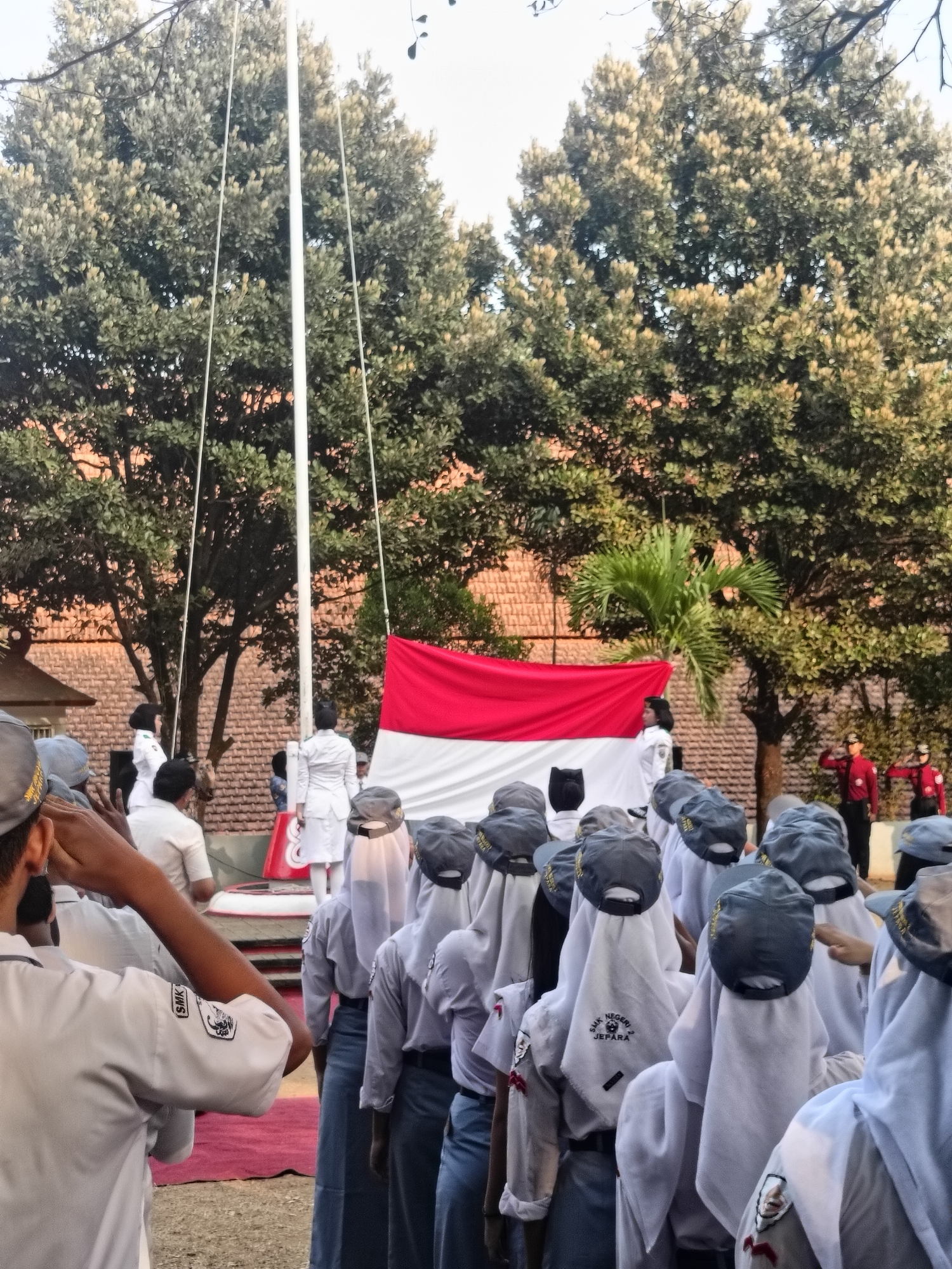 The image depicts an outdoor flag-raising ceremony. In the foreground, a group of students in white shirts, grey hats, and blue skirts are lined up, facing away from the camera. The student in the closest foreground on the left is saluting. Behind them, on a raised platform, several individuals are present. One individual in a white uniform stands next to a flagpole with a large Indonesian flag (red and white) partially unfurled. Two individuals in red uniforms are visible in the background to the right. The environment appears to be a school or public space, with trees and a brick building visible in the background. The lighting suggests it is either early morning or late afternoon.
