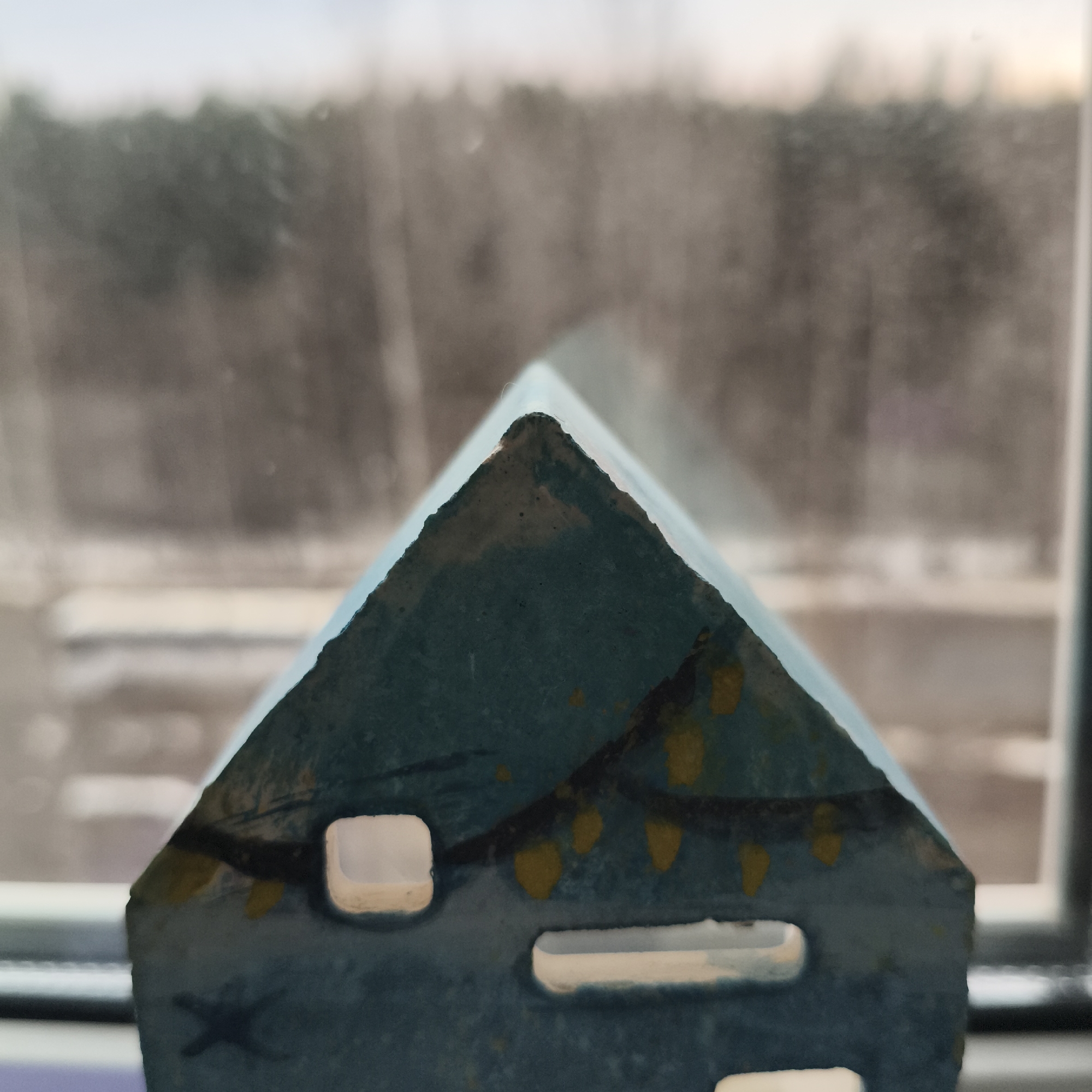A close-up photograph shows a blue, decorative house placed in front of a window. The house is a simple, triangular shape with cut-out window openings. Yellow markings, possibly representing lights or decoration, are painted on the house. The background is blurred, showing what appears to be trees and buildings outside the window. The location is Unknown, Unknown.
