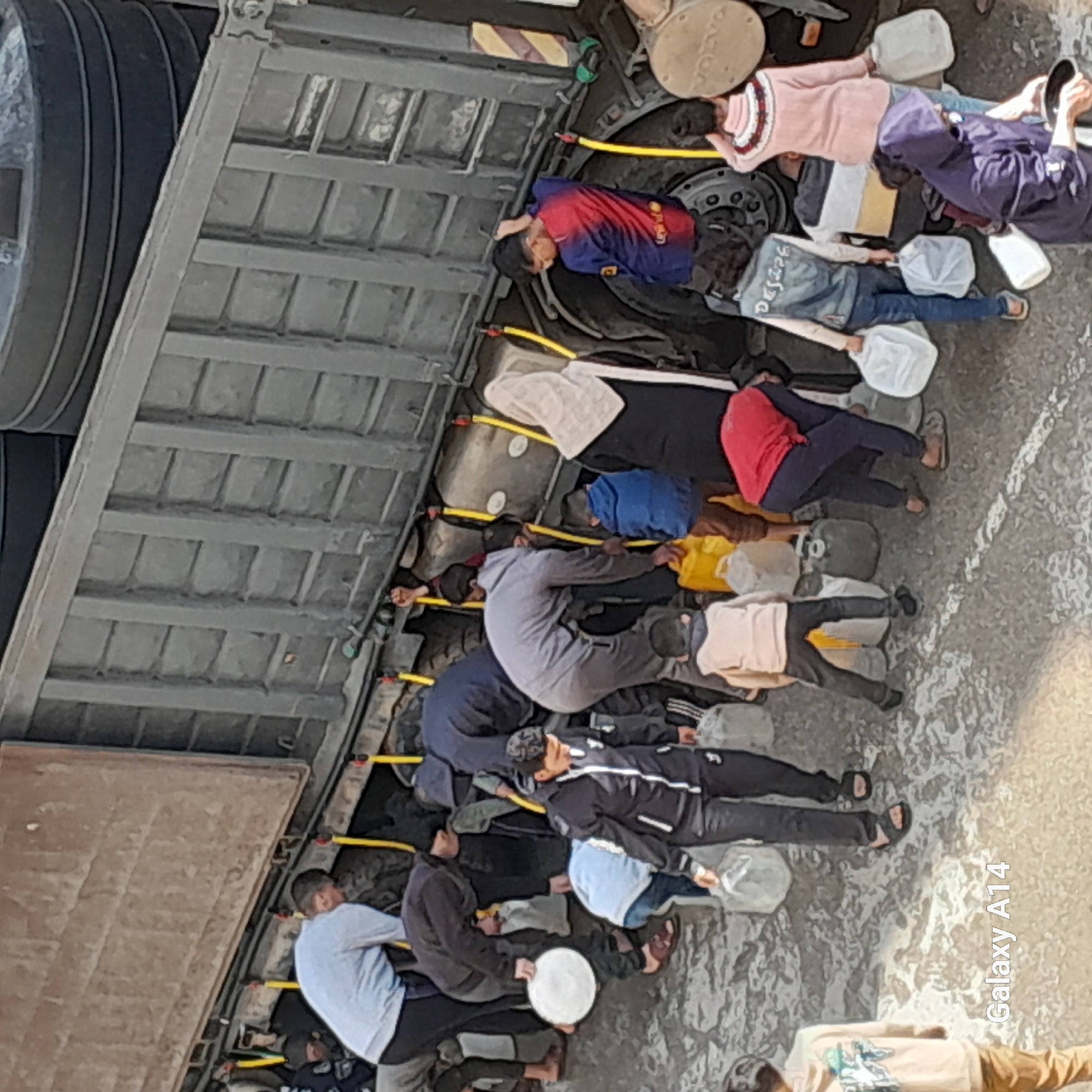 The image depicts a scene where approximately 12 individuals are lying down side-by-side, possibly within or beneath a large vehicle or structure resembling a truck trailer. Some of them seem to be holding white, circular objects or containers. The location is unknown, Unknown.