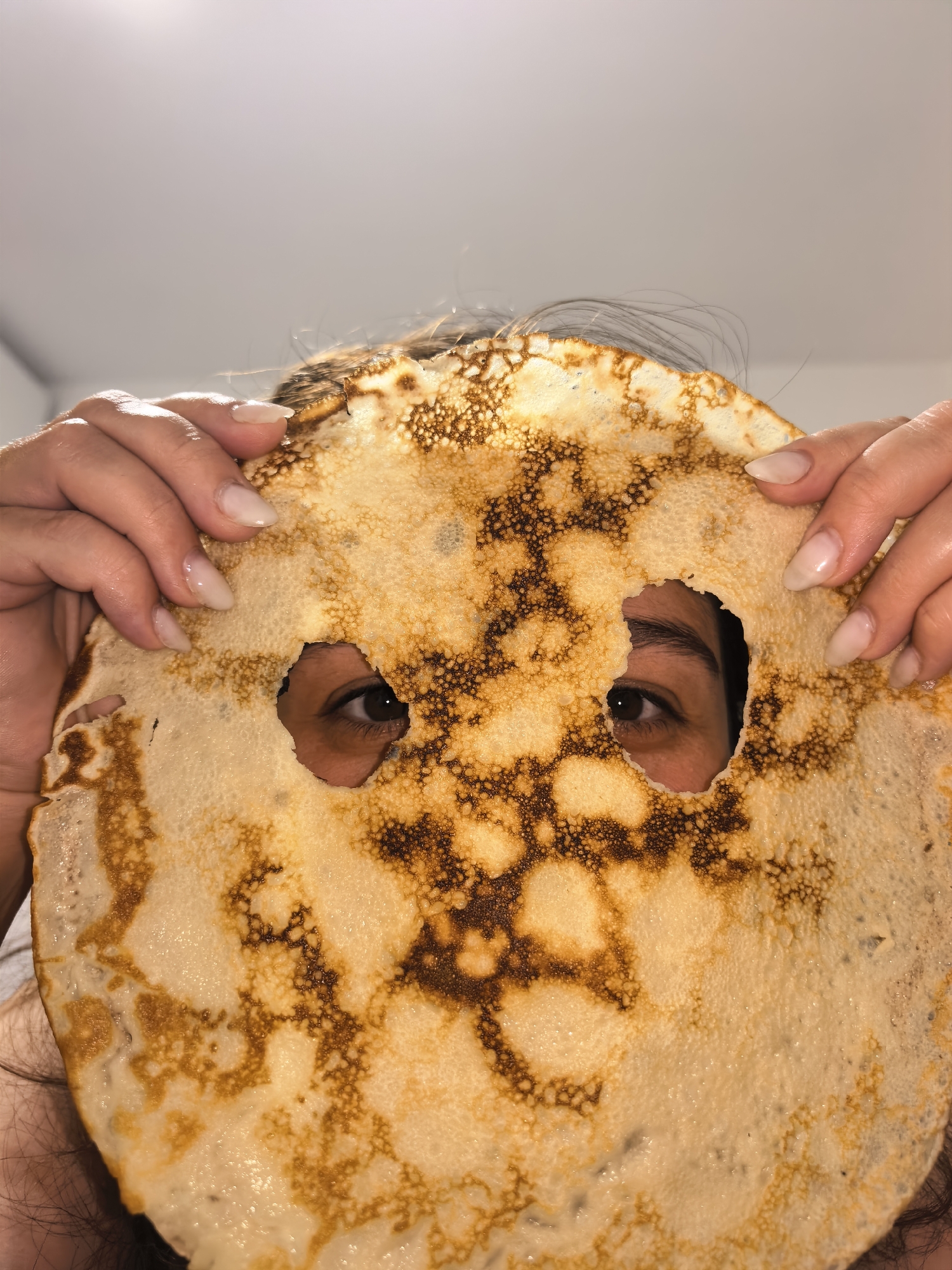 A person is seen holding a large, round pancake in front of their face, using it as a makeshift mask. Two irregular holes are cut into the pancake, allowing the person's dark eyes to be visible through them. The pancake itself is golden-brown with varying shades of darker, cooked areas, suggesting a crêpe or thin pancake. Its surface shows a bubbly texture.
Both of the person's hands are holding the edges of the pancake. Their fingernails are long, almond-shaped, and appear to have a natural or light nude polish. Dark hair is visible peeking out from behind and above the pancake. The background is a plain, light-colored surface, possibly a ceiling or wall, indicating an indoor setting. The lighting is bright and even. No specific emotions are discernible beyond the playful nature of the act. There is no visible text in the image.