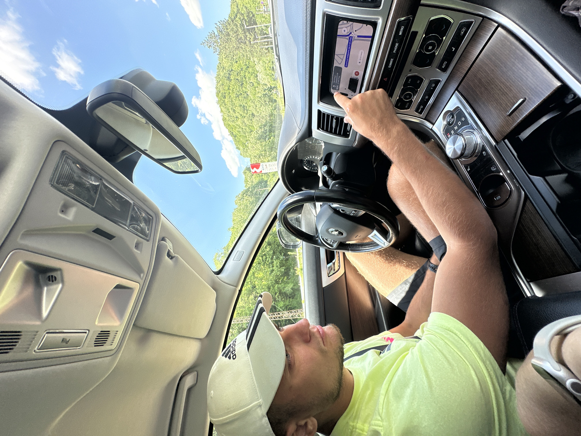 The image shows a young man driving a car, likely a Jaguar, through a scenic, hilly area with lush green trees. He is wearing a white Adidas baseball cap and a neon yellow t-shirt. His right hand is touching the car's touchscreen navigation system, which displays a map. The interior of the car features wood-grain trim and a modern dashboard. The exterior environment suggests a sunny day with a clear blue sky and some white clouds. While the specific location is not visible in the image, the context of Crkvine, Montenegro, implies a mountainous or coastal region with significant natural beauty, which is consistent with the backdrop.