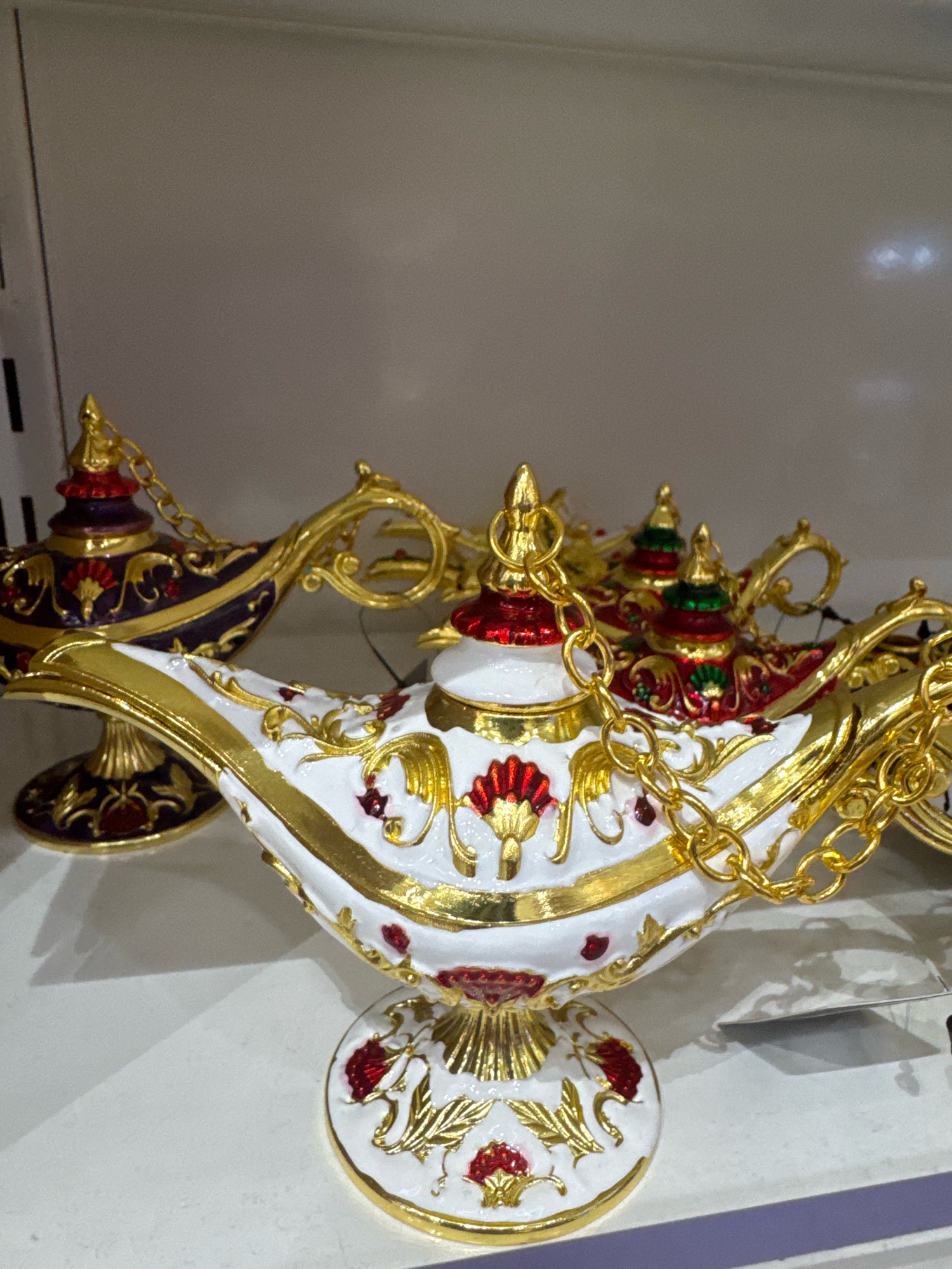 The image shows a close-up of several decorative genie lamps, similar in style to Aladdin's magic lamp. The lamps are ornate, featuring a white base with intricate gold detailing and accents of red and green. One lamp in the foreground is predominantly white with gold scrollwork and red floral motifs. A gold chain connects the lid to the body of the lamp. Behind it, a second lamp is visible, this one appears to be dark purple with gold and red embellishments. Both lamps are displayed on a white shelf, possibly in a retail setting. The background is a plain, light-colored wall. No people are present in the image. The location is stated to be Al Matar, United Arab Emirates, suggesting this might be a souvenir shop or a decorative items store in that area.