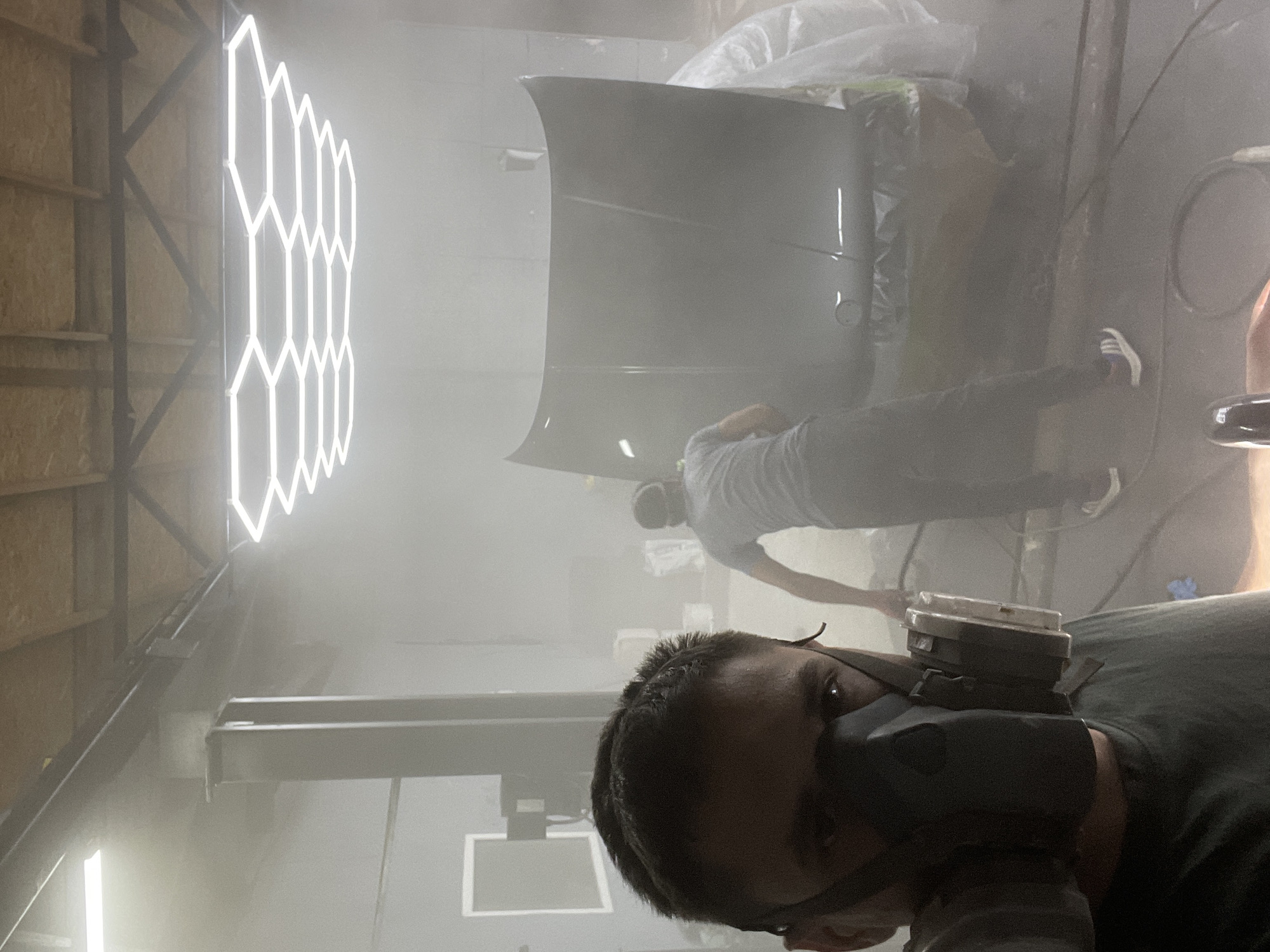 In an indoor workshop, possibly a car repair shop, two individuals are visible amidst a misty atmosphere, likely from a spray painting process. In the foreground, a person is captured in a close-up selfie, wearing a black respirator mask and a dark t-shirt. Their face is partially obscured by the mask, but their eyes are visible, looking towards the camera. In the mid-ground, another individual, wearing a grey t-shirt and dark pants with athletic shoes, is bent over an open car hood, which is the central subject of the image. The hood appears to be undergoing a painting or refinishing process, with a sheen suggesting a wet finish. The environment is characterized by the presence of spray mist, which softens the background details, including industrial lighting fixtures overhead and what appears to be a car lift. The overall scene suggests an active work environment related to automotive bodywork or painting.
