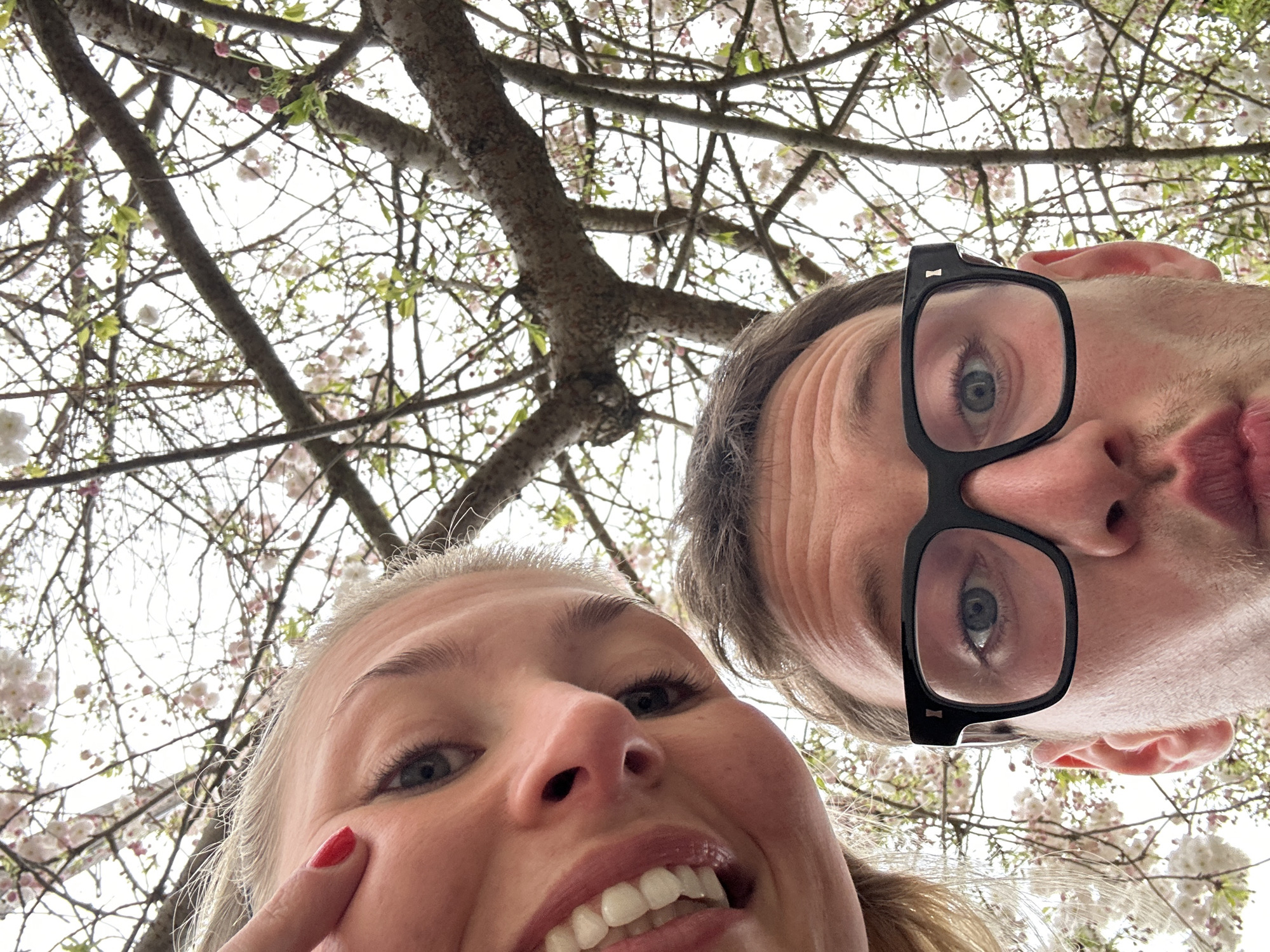 A selfie taken from a low angle features two individuals under a blossoming tree. The woman on the left has blonde hair, red nail polish, and is smiling. The man on the right wears black-framed glasses and appears to be making a puckered expression. The background shows tree branches with pale pink and white blossoms against a bright sky. The location is likely a park in London during springtime due to the presence of flowering trees commonly found in the city's green spaces.