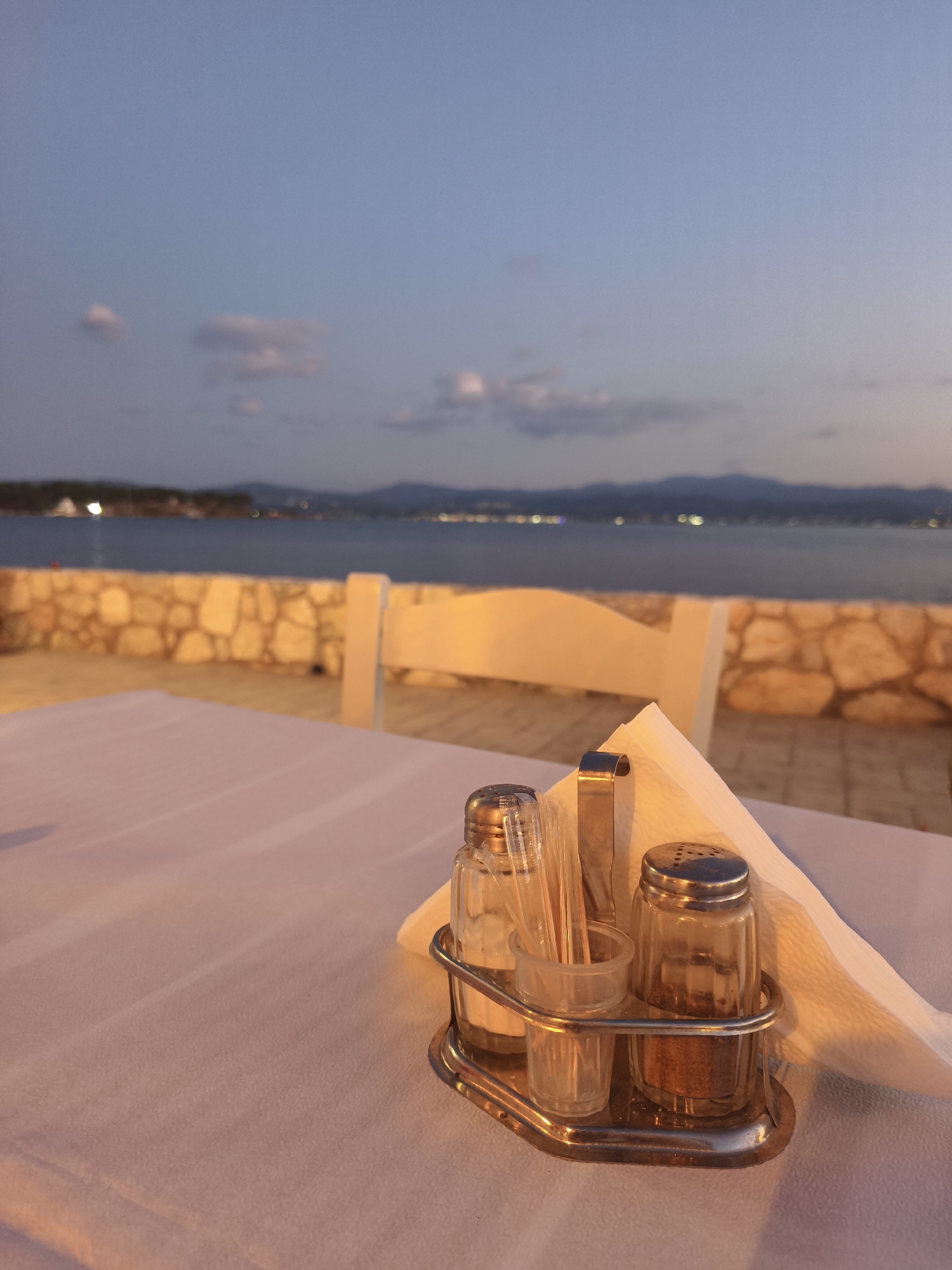 A close-up shot focuses on a table setting at what appears to be a seaside restaurant in the city of Eretria Municipal Unit, Greece, during twilight. The foreground features a white tablecloth adorned with a metal caddy holding a salt shaker, a pepper shaker, toothpicks in a small clear glass holder, and a napkin. Beyond the table, a stone wall separates the dining area from a view of a calm body of water and distant mountains. The sky is a dusky blue with scattered clouds, and lights from the far shore are visible, suggesting a town or village across the water. A white chair is partially visible behind the table. The lighting suggests it is either early morning or late evening, with a warm glow from artificial sources illuminating the wall and parts of the table.