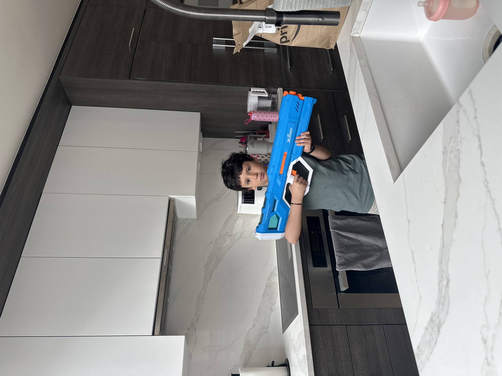 A child is holding a blue X-Shot water gun in a kitchen. The kitchen features white cabinets, a white marble countertop with a sink, a stainless steel oven, and gray cabinets. A vacuum cleaner hose and an Amazon delivery bag are visible above the countertop. The image does not provide enough information to confirm that this scene is in New York.