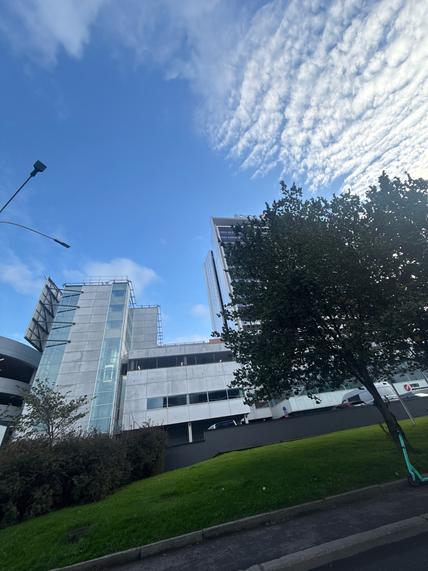 The image shows a modern office building with a glass facade and concrete elements, partially obscured by a large tree in the foreground. The building appears to be located in an urban environment, with a street and sidewalk visible in the lower part of the frame. The sky is blue with wispy white clouds. A street lamp is visible on the left side of the image. On the building's facade, the word "SÕLTUMATU" is visible. The building is located in the Kristiine district of Tallinn, Estonia. A teal electric scooter is parked near the tree on the right side of the image.