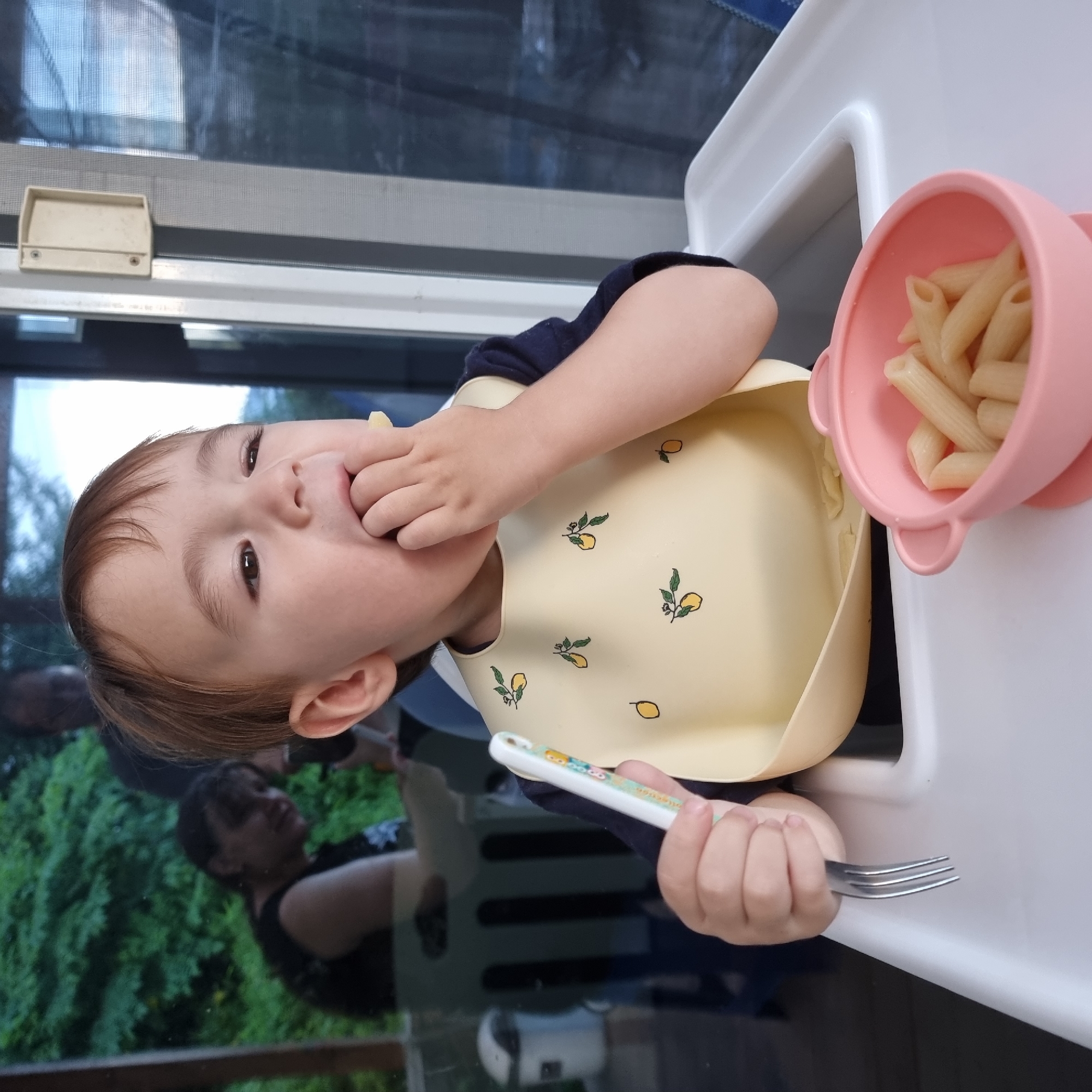 A toddler with brown hair is sitting in a white high chair, eating penne pasta from a pink bowl. The toddler is wearing a yellow bib with lemon patterns. The toddler's right hand is in their mouth, and the toddler is holding a fork in their left hand. The high chair is positioned near a window with a view of greenery and a reflection of people. The scene appears to be indoors, possibly a home, in Dunnae-myeon, South Korea.