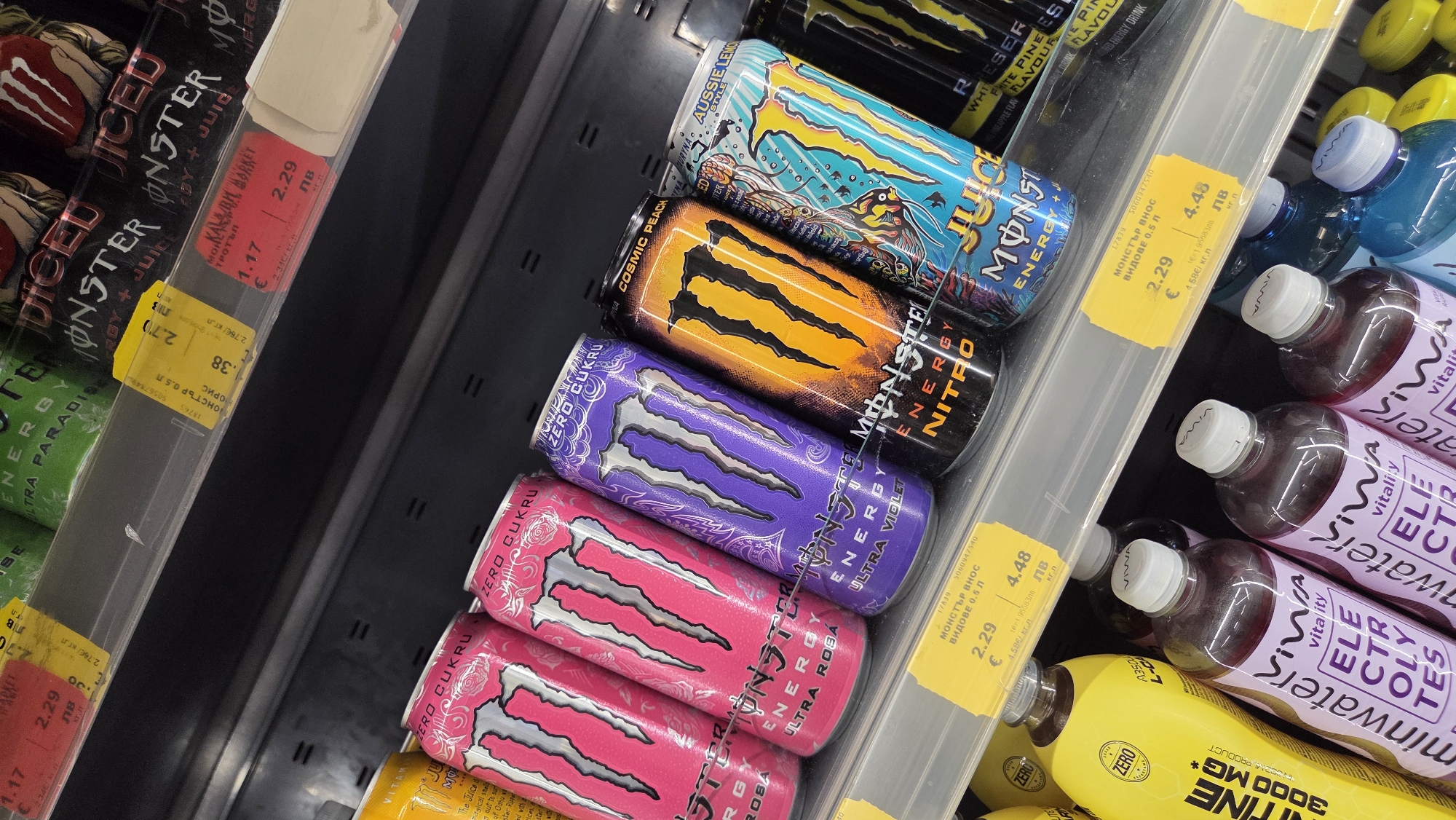 The image shows a shelf in a supermarket in Burgas, Bulgaria, displaying a variety of energy drinks and bottled water. The products are arranged on multiple tiers, with some items in the foreground slightly out of focus. Prominently featured are several cans of Monster Energy drinks in various flavors, including pink (Ultra Rosa), purple (Ultra Violet), orange (Nitro), and turquoise (Juice). Yellow price tags are visible on the shelf in front of some of the drinks, with prices indicated in Euros and Bulgarian Leva. To the left of the Monster cans, there are yellow bottles of Vitality brand electrolyte drinks. Further to the left, a portion of a yellow bottle with the text "VITAMINE 3000 MG*" is visible. The overall lighting suggests an indoor retail environment. No people are present in the image.