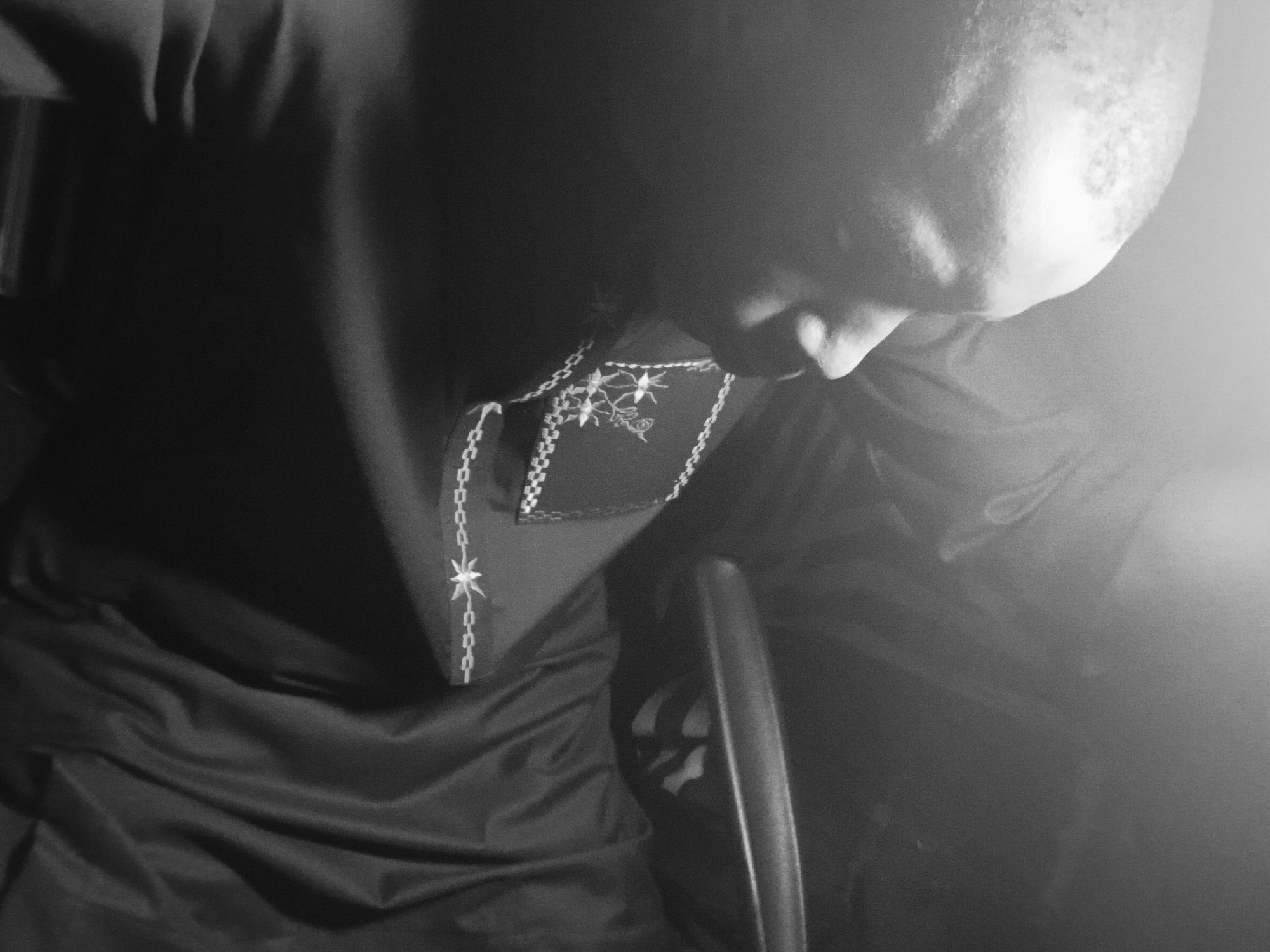 A close-up, high-angle, black and white image shows a person with a shaved head looking down. The person is wearing a dark-colored garment with decorative trim along the neckline and edges. The trim features a chain-like pattern and embroidered star-like shapes. The garment appears to be draped or folded. A portion of a dark object, possibly a chair arm, is visible to the right. The background is dark and obscured by a bright light source. The location is unknown; the image does not provide sufficient information to determine if it was taken in Lagos, Nigeria.