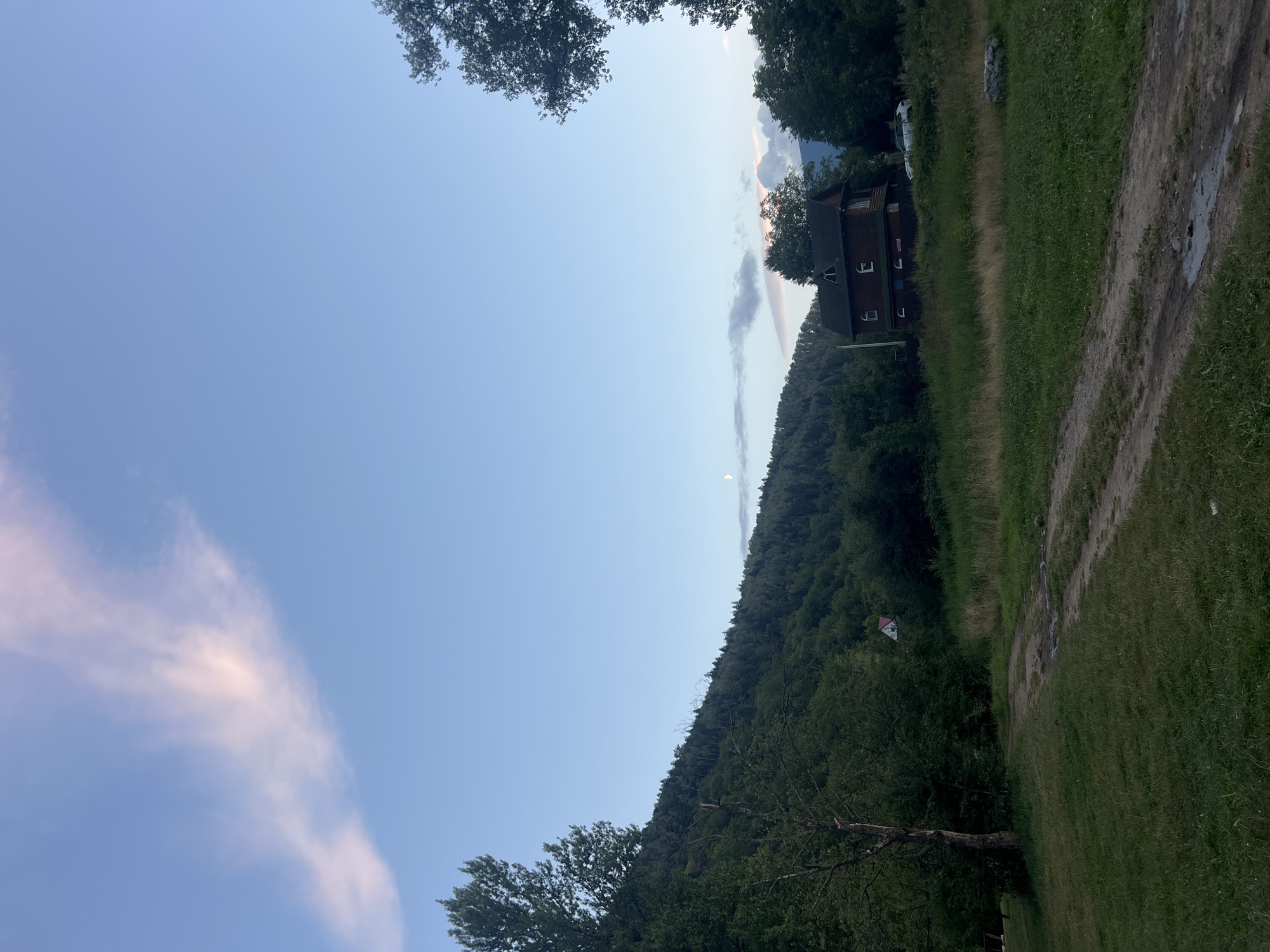 This image depicts a serene, rural landscape likely located in or around Yaremche, Ukraine, characterized by a dirt road meandering through grassy fields, leading towards a dense, forested mountainside. The sky above is a pale blue, transitioning into a softer hue near the horizon, with a delicate wispy cloud formation tinged with pink and a sliver of the moon visible.
Dominating the mid-ground is a traditional wooden house with a dark roof and multiple windows, situated on a slight incline. The architecture suggests a rustic, possibly older, dwelling. A white car is parked near the house, partially obscured by surrounding foliage. The foreground is a grassy area with a dirt path, suggesting it might be a rural road or a pathway leading to the house. The overall impression is one of tranquility and natural beauty, typical of a mountainous region. The vegetation is lush, with tall trees framing the scene on either side, and the mountainside covered in a dense canopy of green trees. The lighting indicates it may be late afternoon or early evening, given the soft quality of light and the presence of the moon and sunset-tinged clouds. There are no people visible in the image, emphasizing the quiet and secluded nature of the location.
