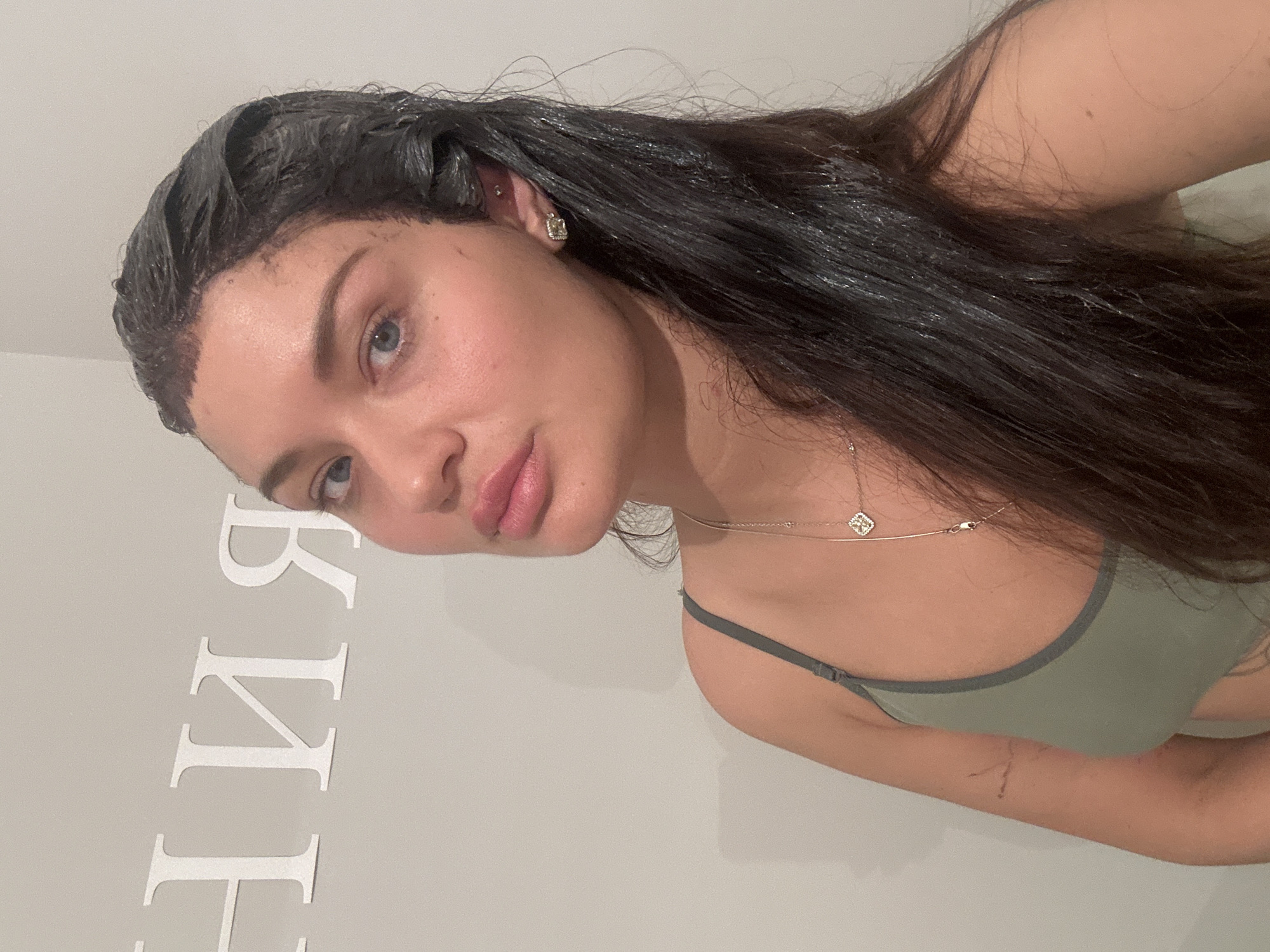 The image is a rotated close-up selfie of a woman indoors, possibly in a personal space. Her head is positioned to the left side of the frame, and her body extends towards the right. She has long, dark hair which appears to be covered in a dark product, likely a hair dye or treatment, visible around her roots and hairline. Her eyes are blue, and she looks directly towards the camera with a neutral expression.
She is wearing an olive-green or dark sage-colored bralette with grey trim. Around her neck, she wears a delicate, gold-toned chain with a small, square-shaped pendant. She has a square stud earring in her right ear and a small additional piercing higher up on the ear.
The background is a plain, light-colored wall. On the lower left section of the wall, several large, white, serif-style letters are affixed. Due to the image's rotation, the letters appear to be oriented on their side. When mentally rotated to an upright position and read from bottom to top, the visible letters spell "ЯНИКЕН" (YANIKEN) in Cyrillic script. No other specific location cues or time of day indicators are visible within the image itself.