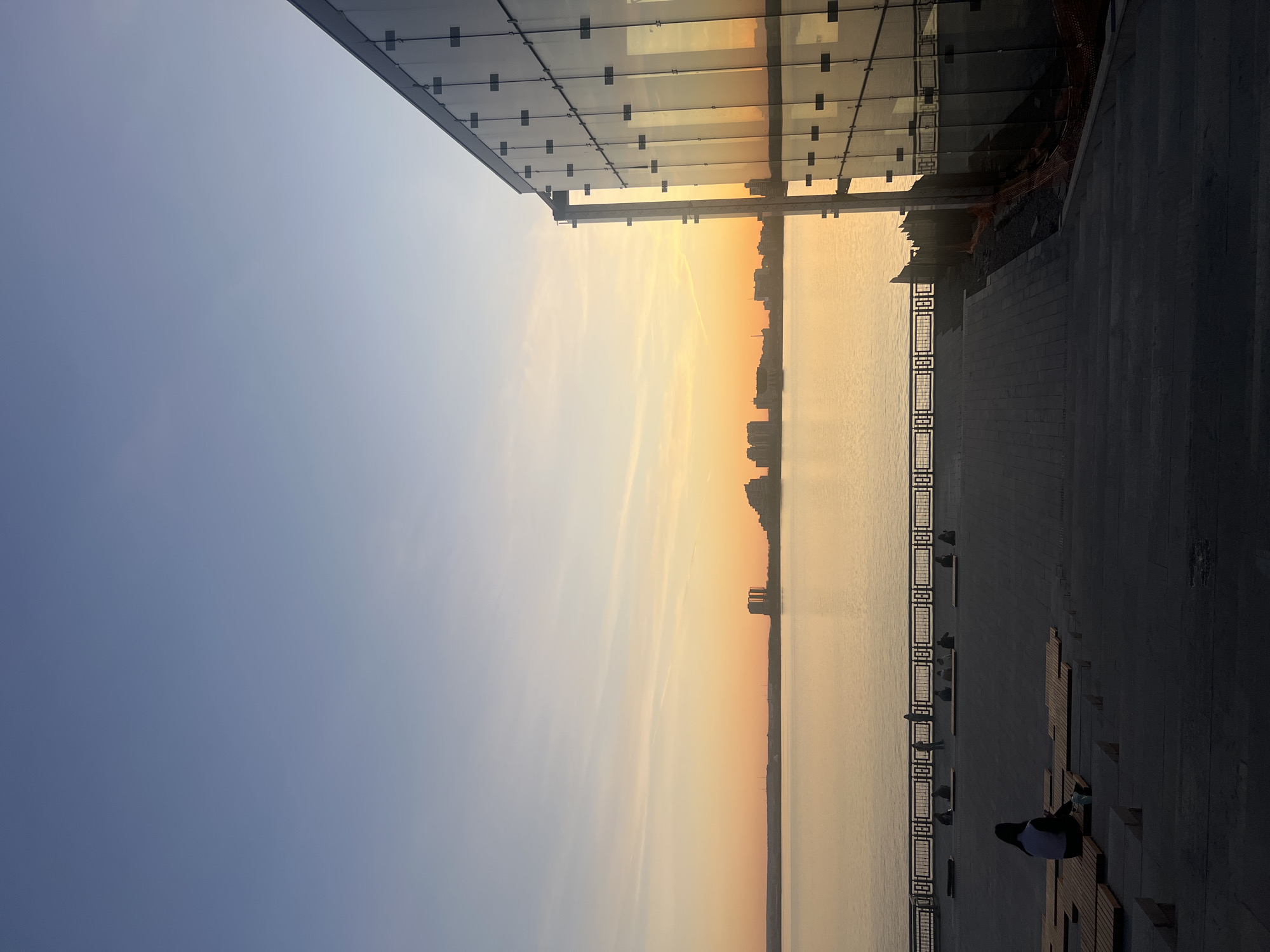 The image captures an expansive urban waterfront scene during sunset or sunrise, evidenced by the warm, golden light painting the sky and reflecting on the water. The sky transitions from a soft blue on the left to vibrant orange and yellow near the horizon on the right.
A wide body of water, likely a river or large lake, dominates the central part of the frame, its surface gently reflecting the sky's colors. Across the water, a distant cityscape forms a dark silhouette against the bright horizon, indicating an urban setting.
On the left side, a modern building with a glass facade is prominent, its panes subtly reflecting the ambient light. Below this building and running along the water's edge is a paved embankment or promenade, featuring decorative metal railings.
Several people are visible along the embankment. Most are indistinct, silhouetted figures, either standing or seated on benches. In the immediate foreground, one person is clearly visible, seated on a unique, abstract-shaped wooden bench. They are dressed in dark attire with a contrasting white section, possibly a top or hoodie, and appear to be looking out towards the water.
The scene conveys a sense of tranquility and leisure, suggesting a popular spot for observing the beautiful sky. Given the context of Kazan, Russia, this could be an embankment along the Kazanka River or Volga River. There is no visible text in the image.
