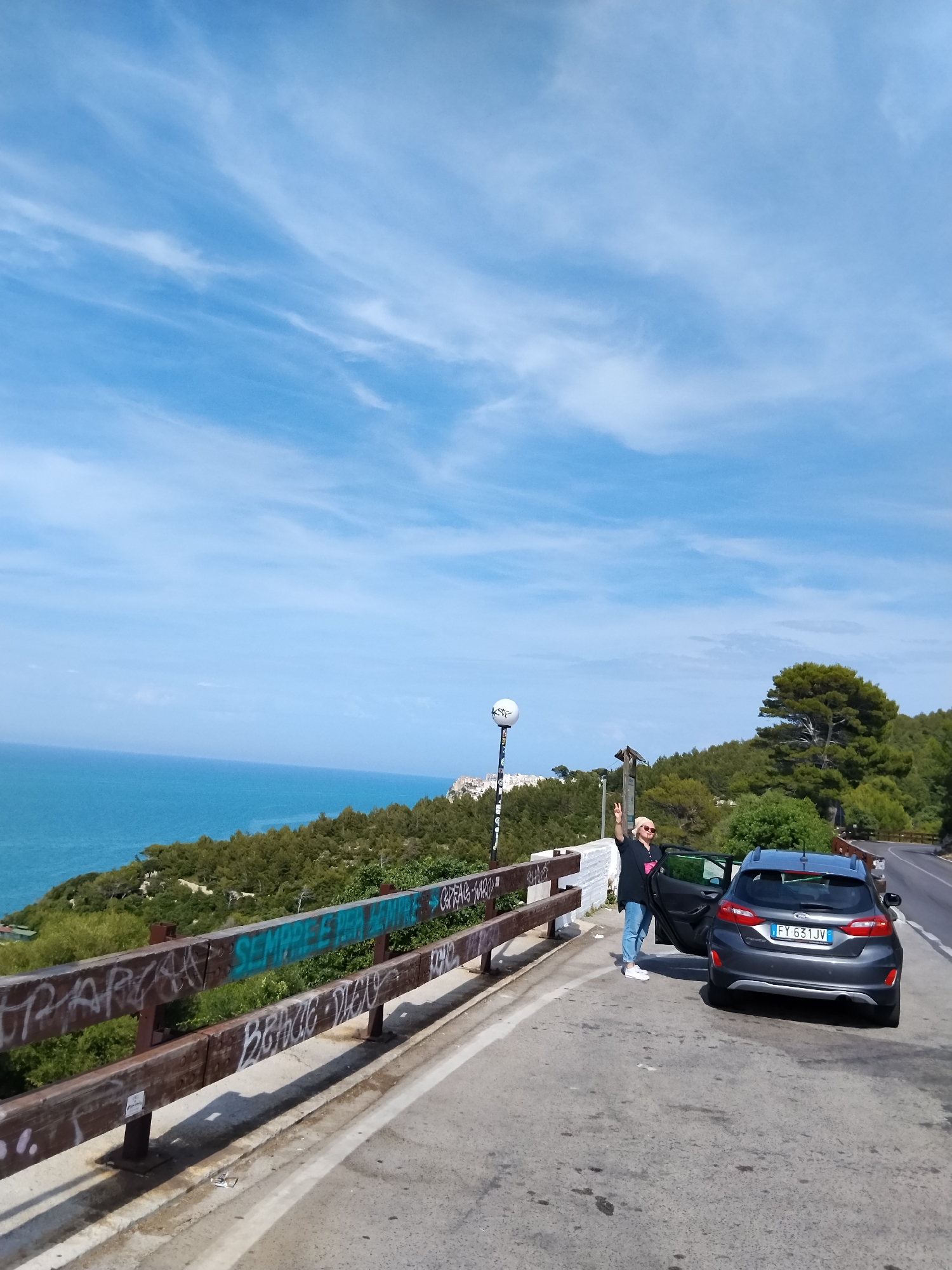 A single individual stands on the paved shoulder of a winding coastal road in Vico del Gargano, Italy. The person, wearing light blue jeans, white sneakers, a dark top, a light-colored baseball cap, and sunglasses, poses with their right arm raised, displaying a peace sign. They stand next to a dark grey Ford Fiesta hatchback, license plate FY 631 JV (Italy), with its driver's side door open. The road curves to the right beyond the car. A wooden and metal guardrail, heavily covered with graffiti including "SEMPRE PER AMORE" in green, separates the road from a steep, forested hillside leading down to the turquoise-blue sea. In the distance, white buildings are visible on a prominent hill to the left. A spherical white light fixture on a black pole stands behind the person. The sky is bright blue with scattered white, wispy clouds. The scene is illuminated by bright daylight.