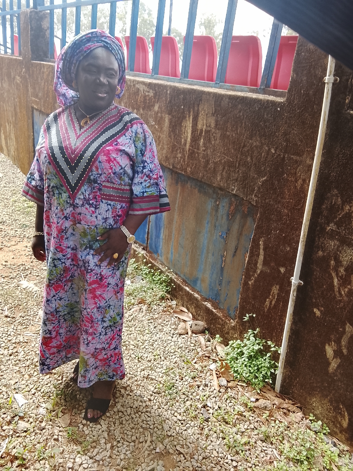 A woman is captured in a full-length portrait, standing outdoors in Zawan, Nigeria. She is smiling broadly and looking towards the camera, conveying a sense of happiness and ease. She is dressed in a vibrant, knee-length kaftan-style garment featuring a tie-dye or batik-like pattern in shades of pink, blue, green, and white. The dress has a prominent V-neckline adorned with horizontal black, grey, and pink stripes, accented with small, light-colored embellishments. A matching patterned headwrap covers her hair.
Her accessories include a distinctive gold-colored pendant necklace, large gold-colored hoop earrings, a delicate gold-colored bracelet on her right wrist, and a silver-toned watch on her left wrist. A large, ornate gold-colored ring is visible on her right ring finger. She wears simple dark sandals.
The woman stands on a light-colored gravel ground, which shows scattered dry leaves and sparse green vegetation. To her right is a tall, rough, dark brown wall, possibly concrete, exhibiting signs of wear and tear with patches of peeling blue paint towards the bottom. A light-colored metal pipe runs vertically against this wall. Above the wall, a dark metal railing is visible, behind which several rows of bright red stadium seats can be seen, suggesting the setting is near a sports facility or arena. The bright, even lighting indicates it is daytime, likely a sunny day. No visible text is present in the image.