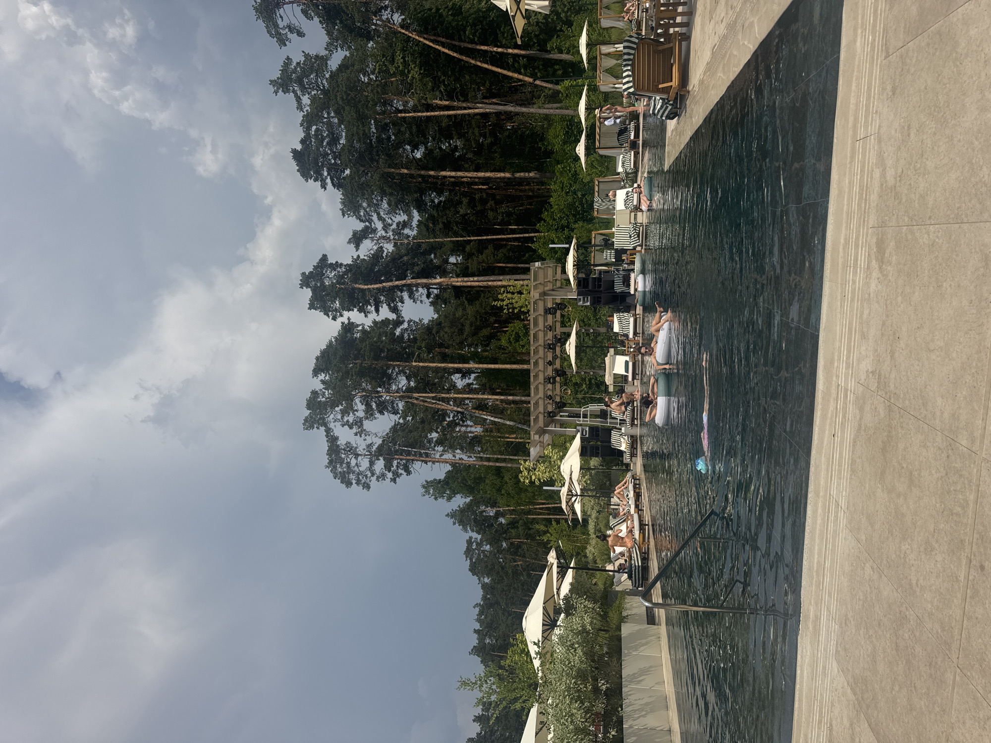 In Razdory, Russia, a resort-style outdoor swimming pool is depicted under a cloudy sky. The pool, filled with dark, rippling water, features a modern, dark tiled bottom and a stone-tiled edge. Several people are present: some are floating on inflatable rings in the water, while others are relaxing on lounge chairs arranged around the pool. The lounge chairs are striped black and white and accompanied by white umbrellas providing shade. In the background, a dense forest of tall pine trees forms a natural backdrop. A wooden structure with speakers and lighting equipment suggests a stage or entertainment area. The overall scene is one of leisure and relaxation in a natural, resort setting.