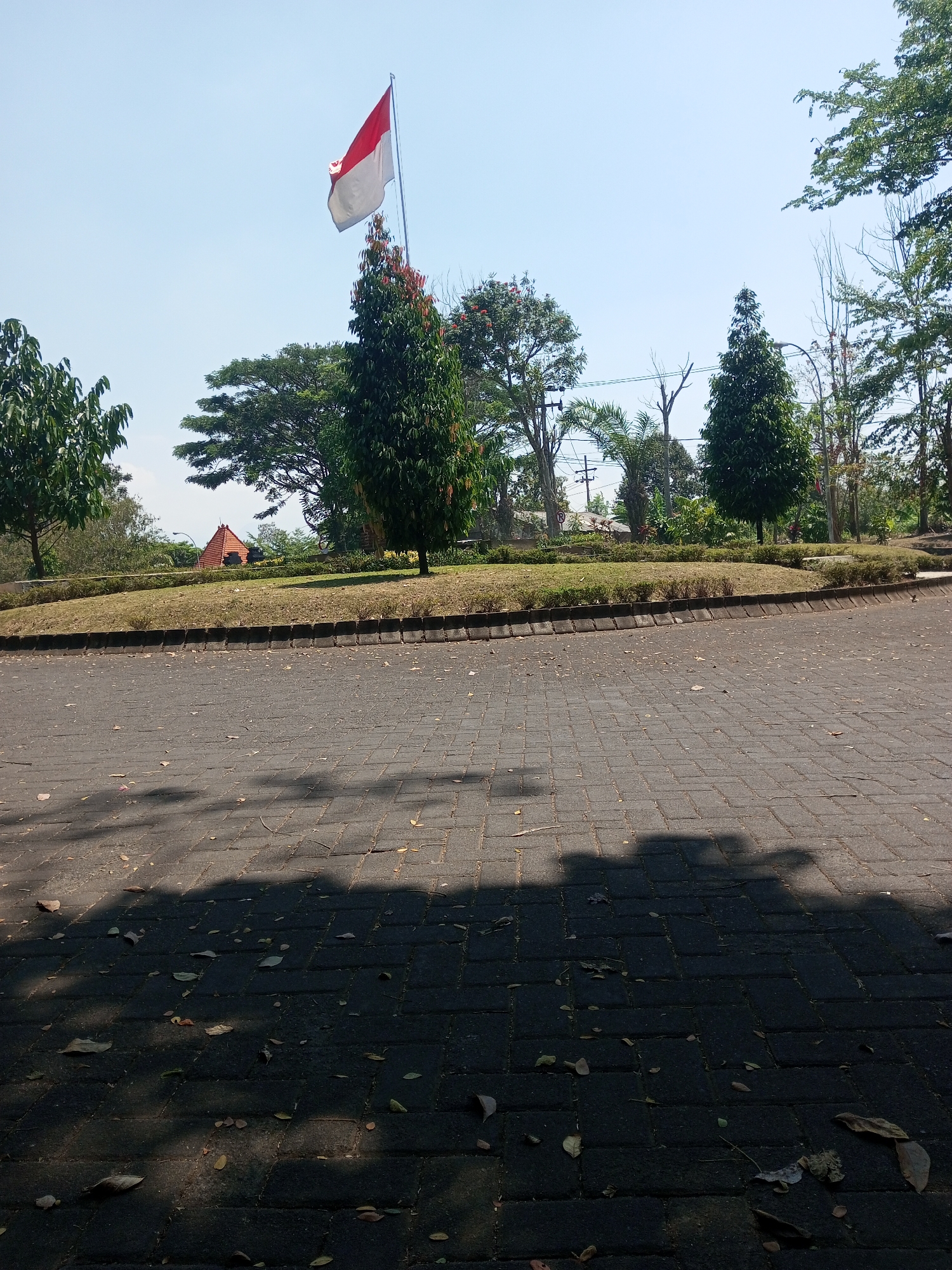 A low-angle, outdoor shot captures a paved area in the city of Klampok, Indonesia, on a bright, sunny day. The pavement is made of dark gray interlocking rectangular blocks, scattered with fallen leaves. Shadows of trees are cast across the surface. In the background, a grassy embankment is lined with low bushes. A red and white Indonesian flag flies from a tall flagpole positioned behind a cluster of trees. Beyond the embankment, a few buildings, including one with a distinctive orange, pyramid-shaped roof, are visible. Several trees of varying sizes and species, including a tall, conical evergreen, populate the scene. The sky is clear and light blue. No people are present in the image.