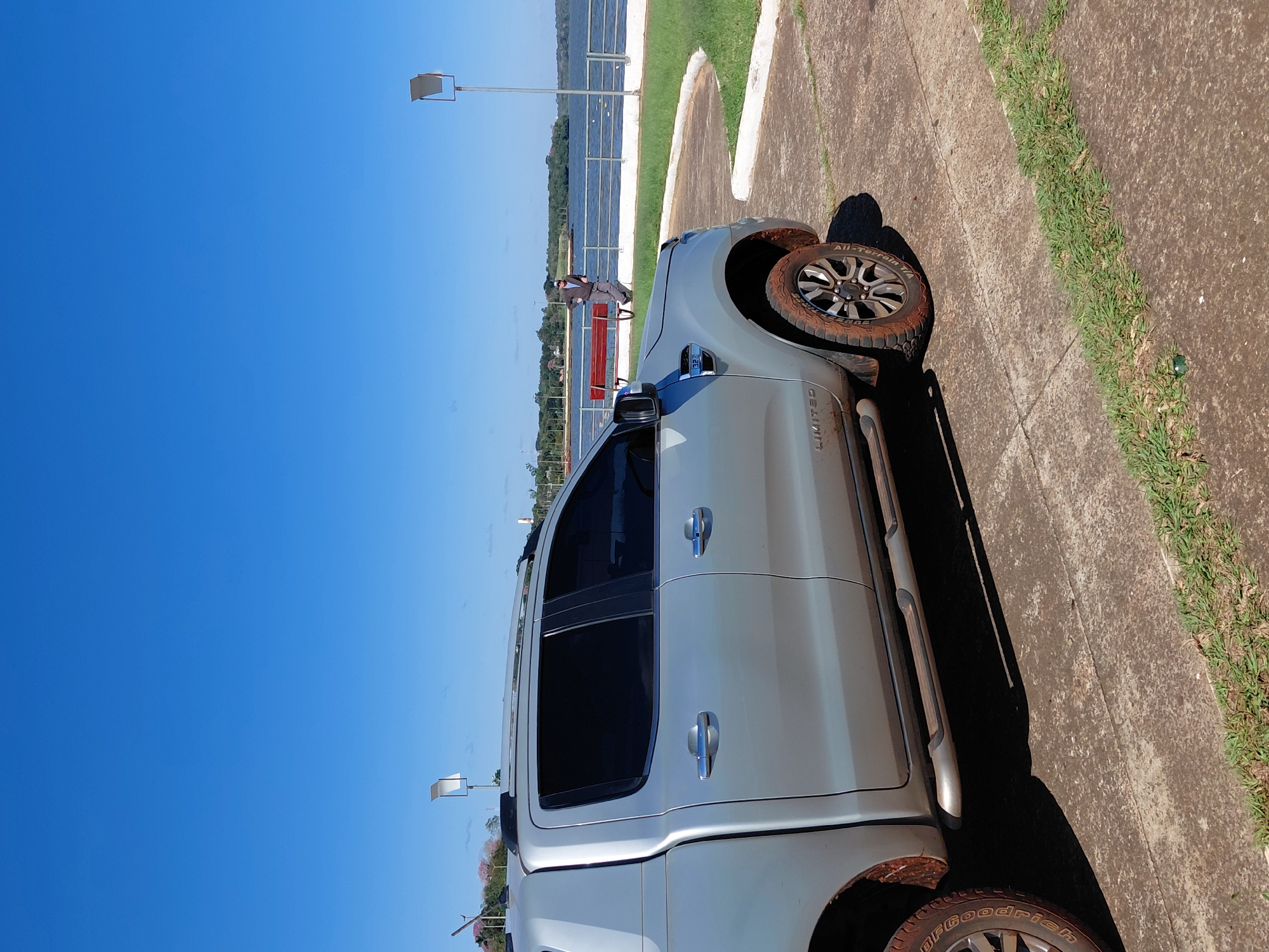 A silver pickup truck is parked on a concrete pathway next to a grassy area. The truck's tires are caked with mud. In the background, a man in a light-colored shirt and dark pants stands near a red bench by a body of water. The sky is clear and blue. This scene is likely located at the Costanera de Encarnación, Paraguay, a popular waterfront promenade.