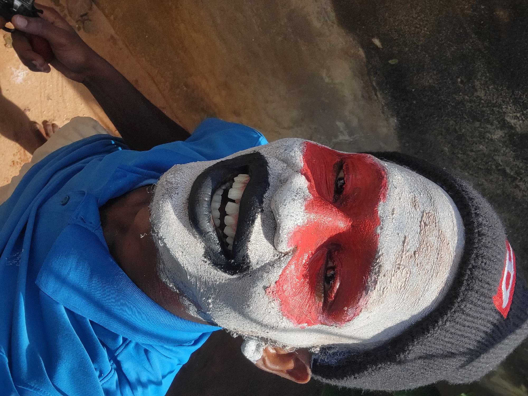 A person with white face paint and a red stripe over the eyes and a black painted mouth is shown. The person is wearing a blue collared shirt, a black knit hat with a red and white emblem, and holding a black object. The background is a light brown wall with some dark patches.